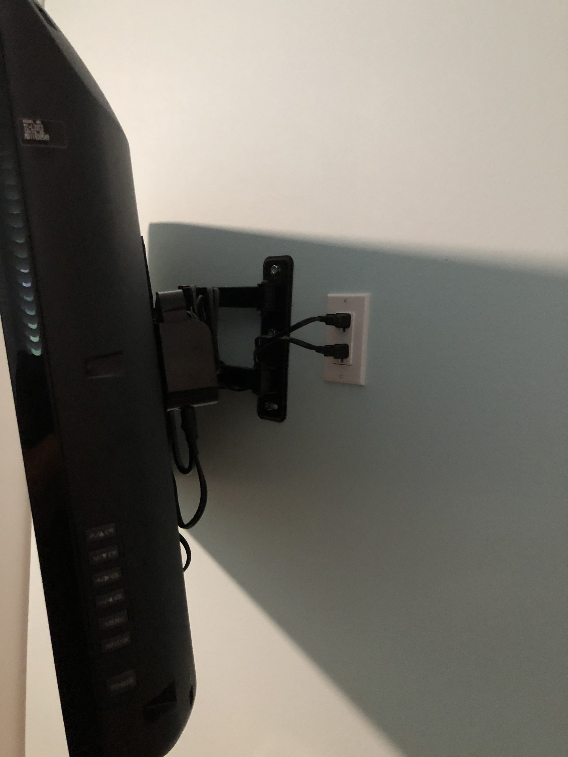 A flat-screen TV is mounted on a wall with a black adjustable bracket, showcasing a neat installation. Several cords are plugged into an outlet behind the TV, and the light blue wall is visible in the background. , Thousand Oaks Handyman, Westlake Handyman, Oak Park Handyman, Agoura Handyman