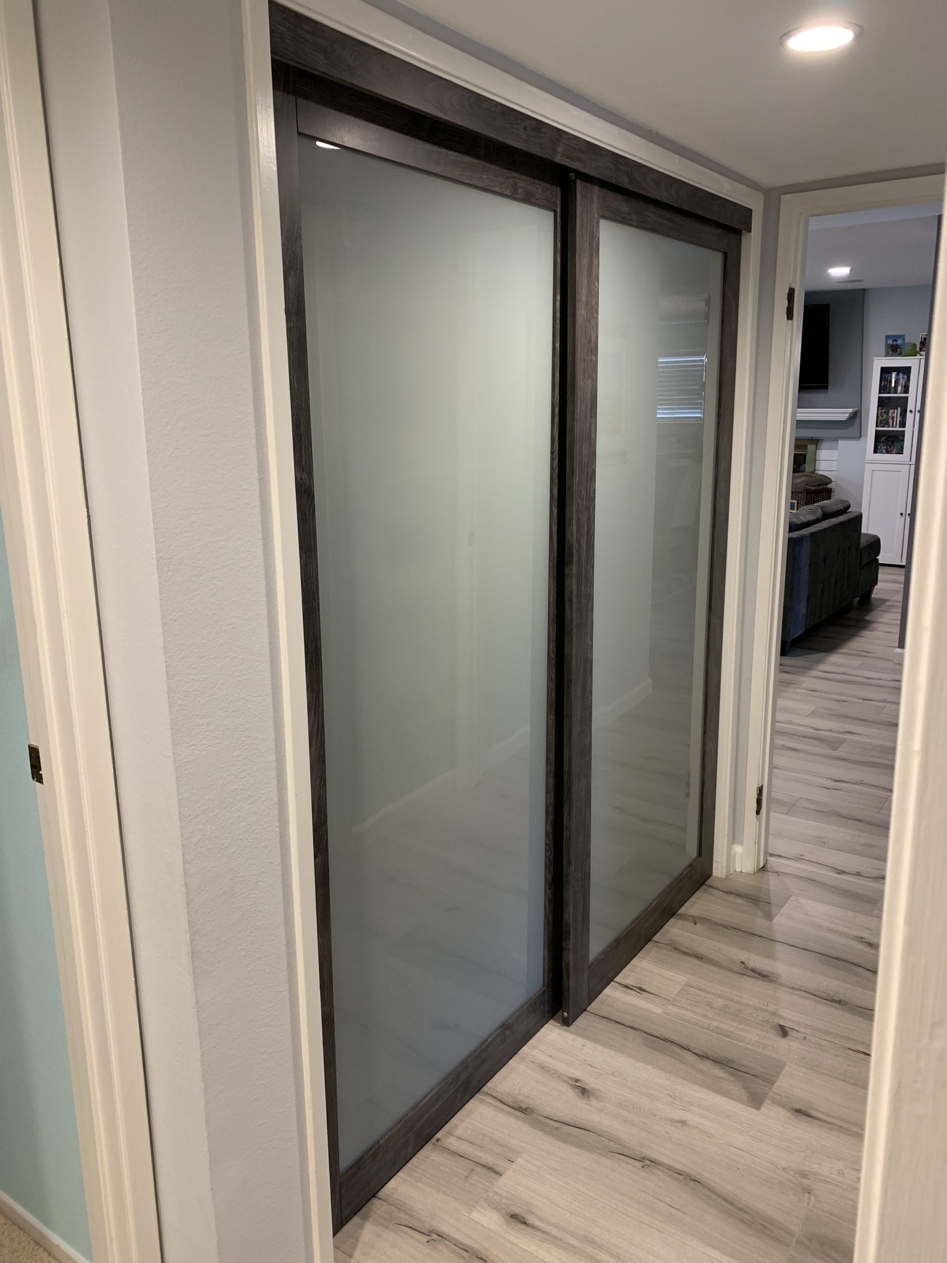 Reliable installation of frosted glass sliding closet doors with dark wood frames enhances the hallway’s light-colored walls and wood-patterned flooring. A living area is visible in the background. , Thousand Oaks Handyman, Westlake Handyman, Oak Park Handyman, Agoura Handyman