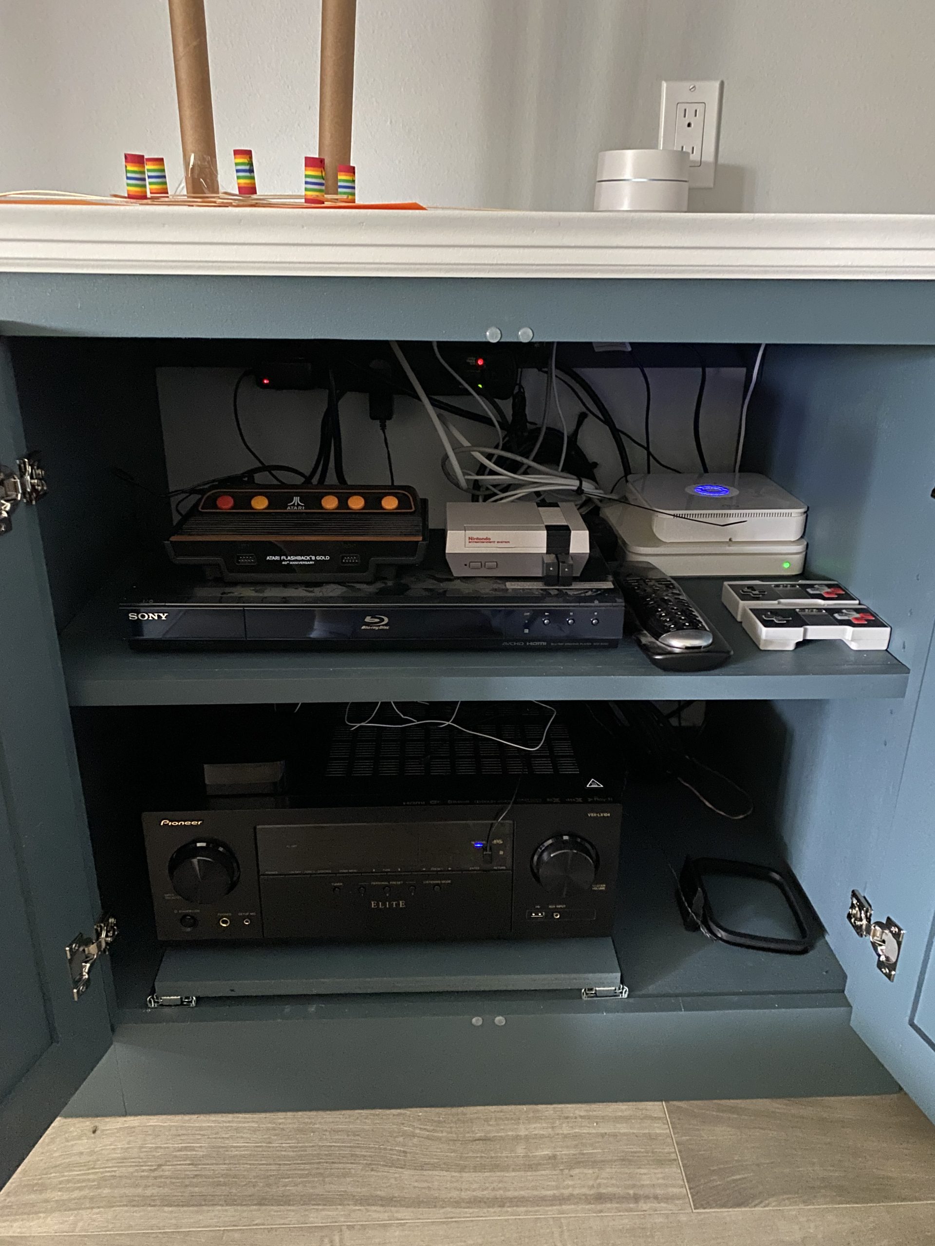 An open blue cabinet holds a Sony Blu-ray player, Pioneer Elite receiver, retro game consoles, remotes, and tangled wiring beneath the shelf inside.