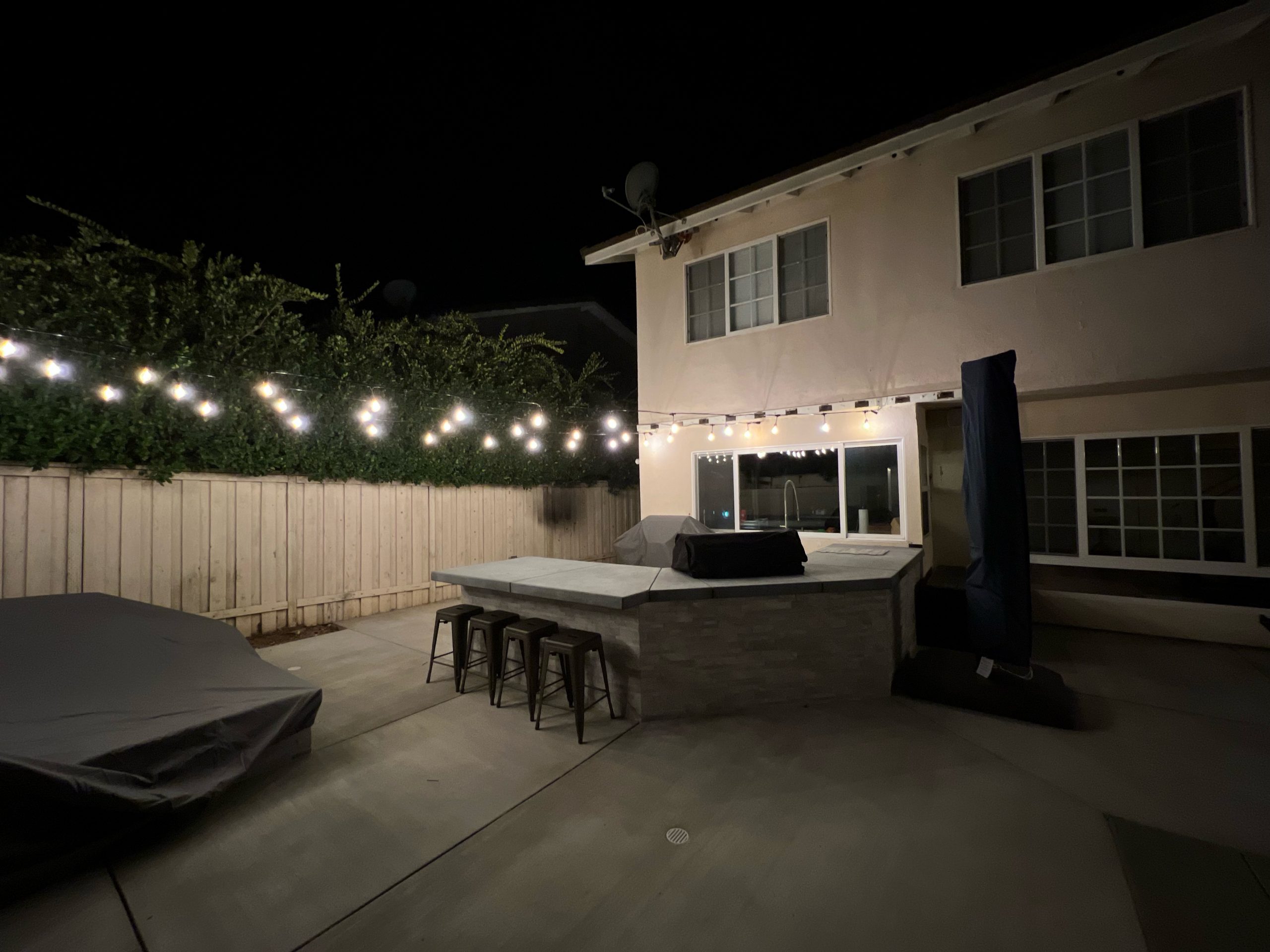 Warm bistro string lights glow across a backyard patio at night beside a two-story house, covered furniture, and a wooden fence under a dark sky.