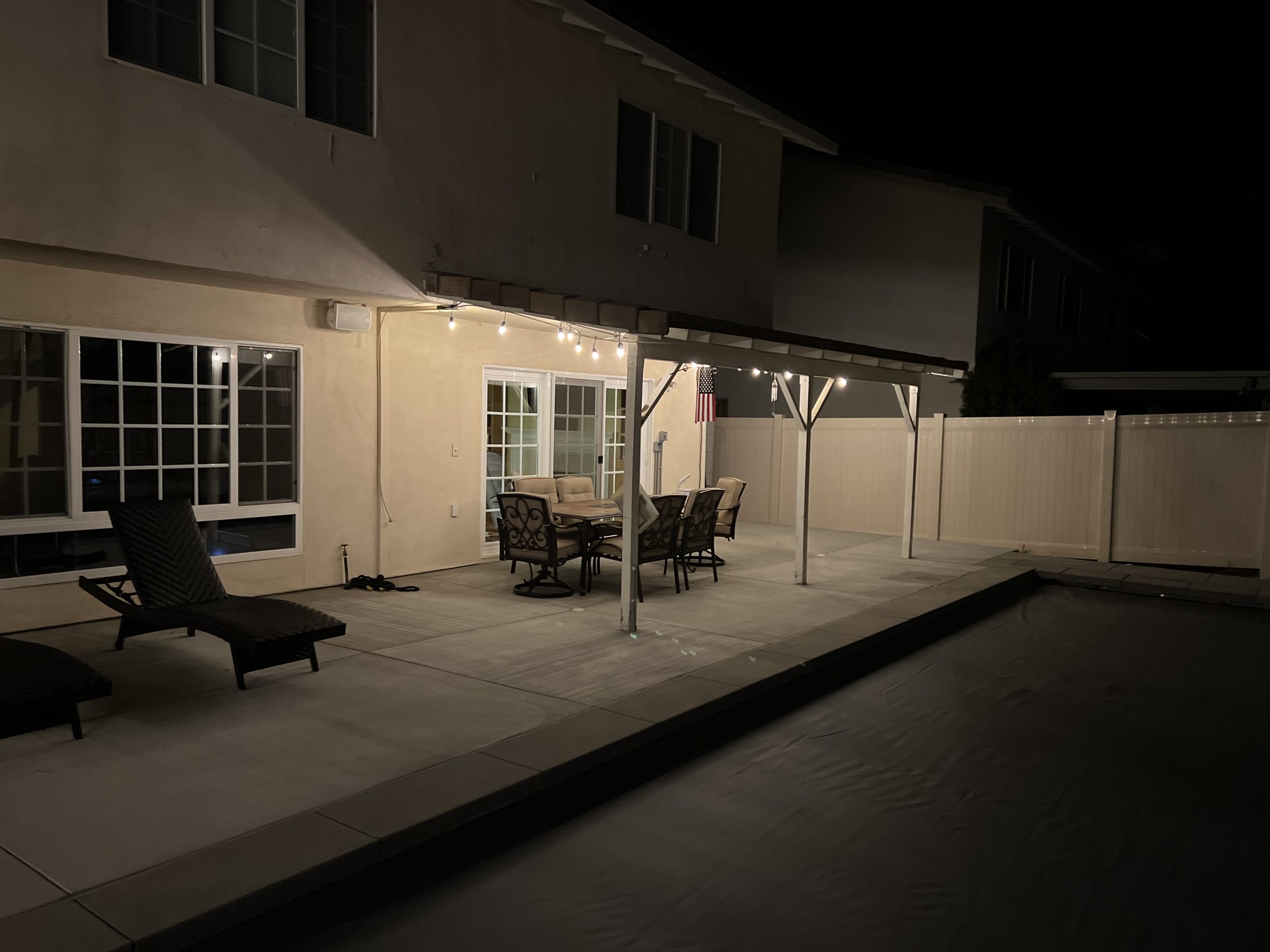 A lit backyard patio with string lights under the awning, a dining set by sliding doors, and lounge chairs beside a dark pool at night.