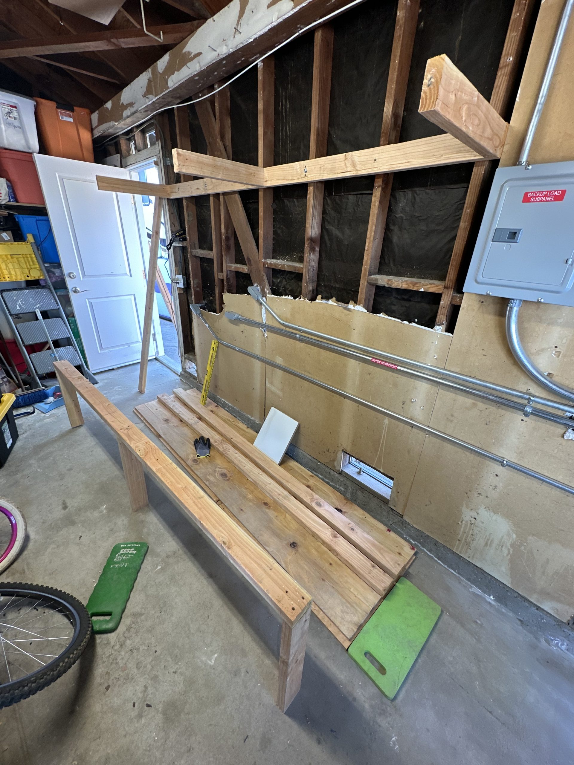Unfinished wooden shelving frame spans a garage wall beside exposed studs, electrical panels, stored bins, and loose boards on the floor during construction.