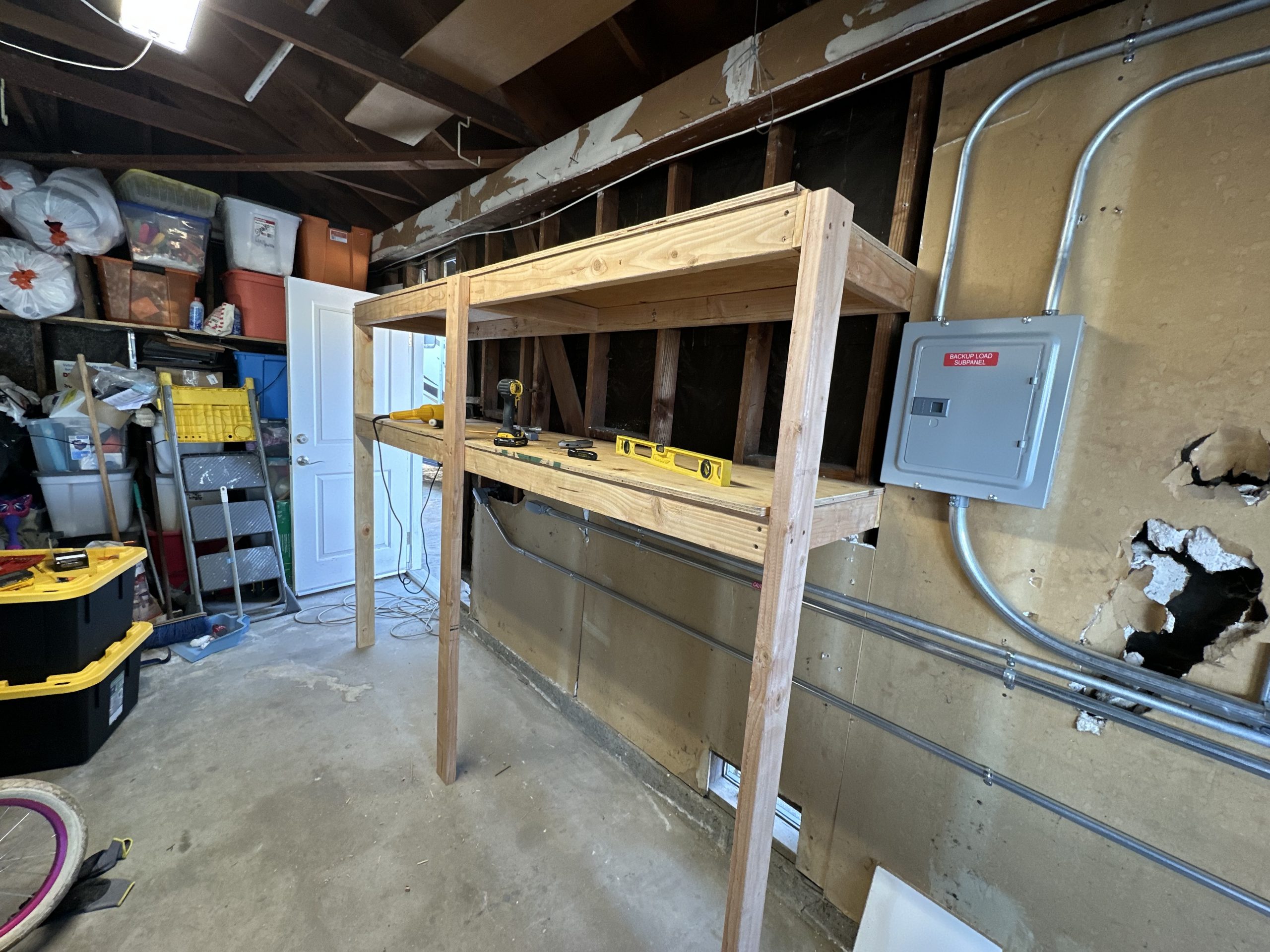 An unfinished wooden shelving frame stands in a cluttered garage workspace beside storage bins, hand tools, exposed wiring, and a gray subpanel.