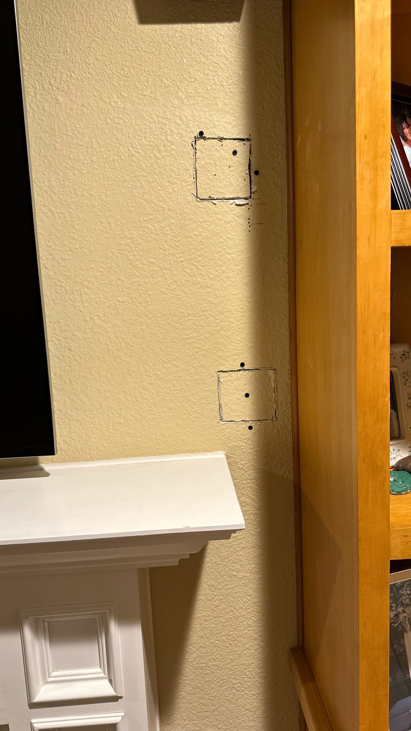Two square drywall patches and small screw holes mark a beige textured wall beside a wooden cabinet and white mantel in warm indoor light.