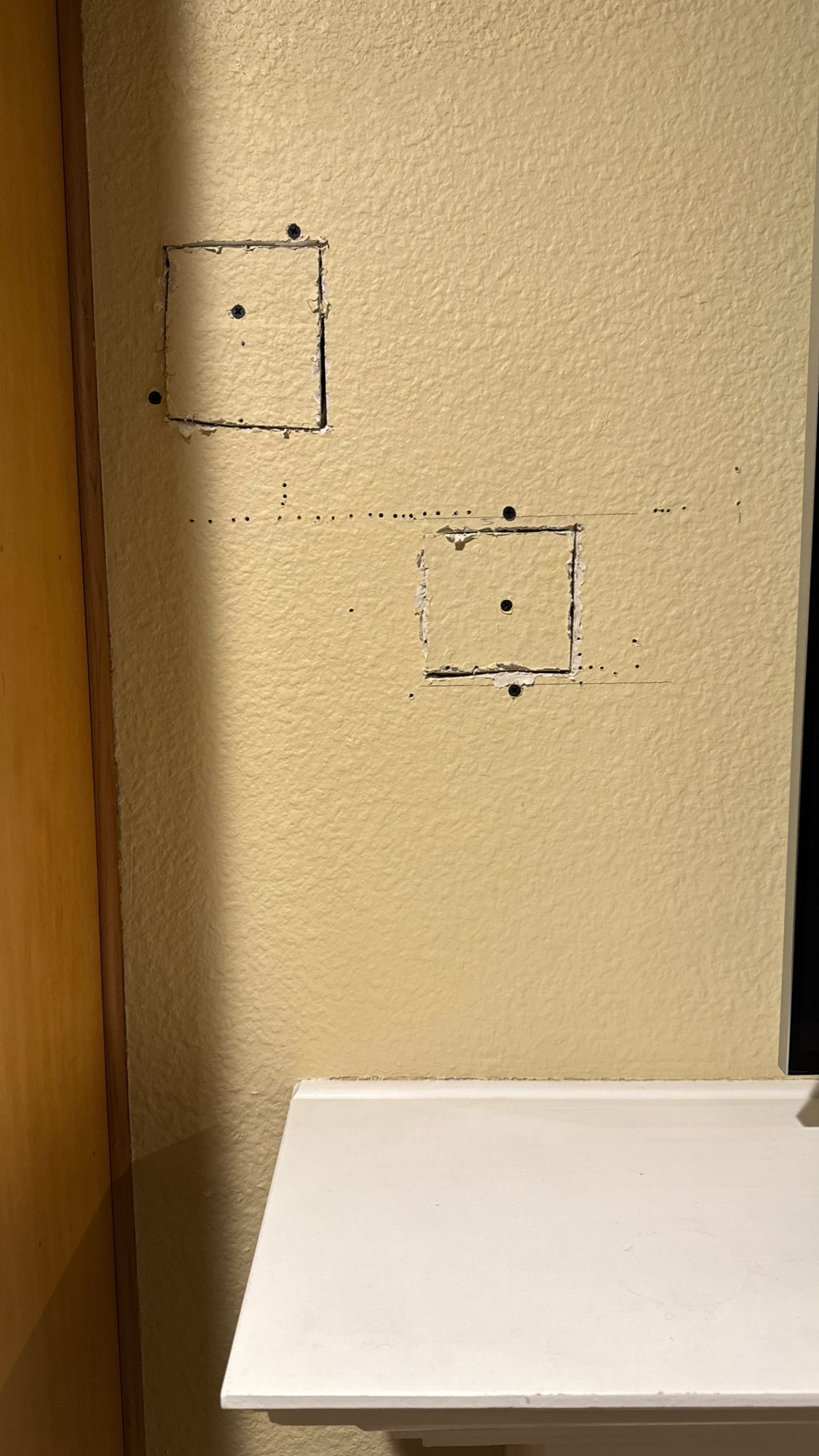 Beige interior wall with two square drywall patches, scattered nail holes, and unfinished seams beside wooden trim and a white shelf at right.