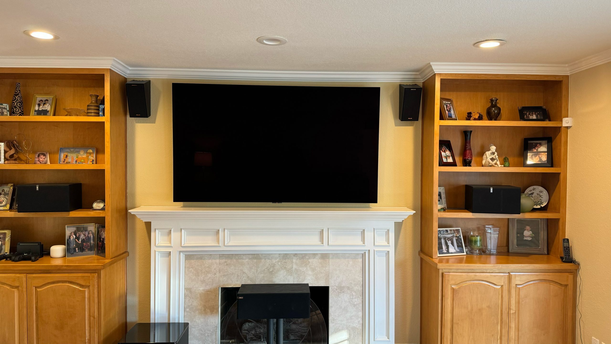 A large black TV hangs above a white fireplace, flanked by built-in wooden shelves filled with framed family photos, vases, and black speakers.