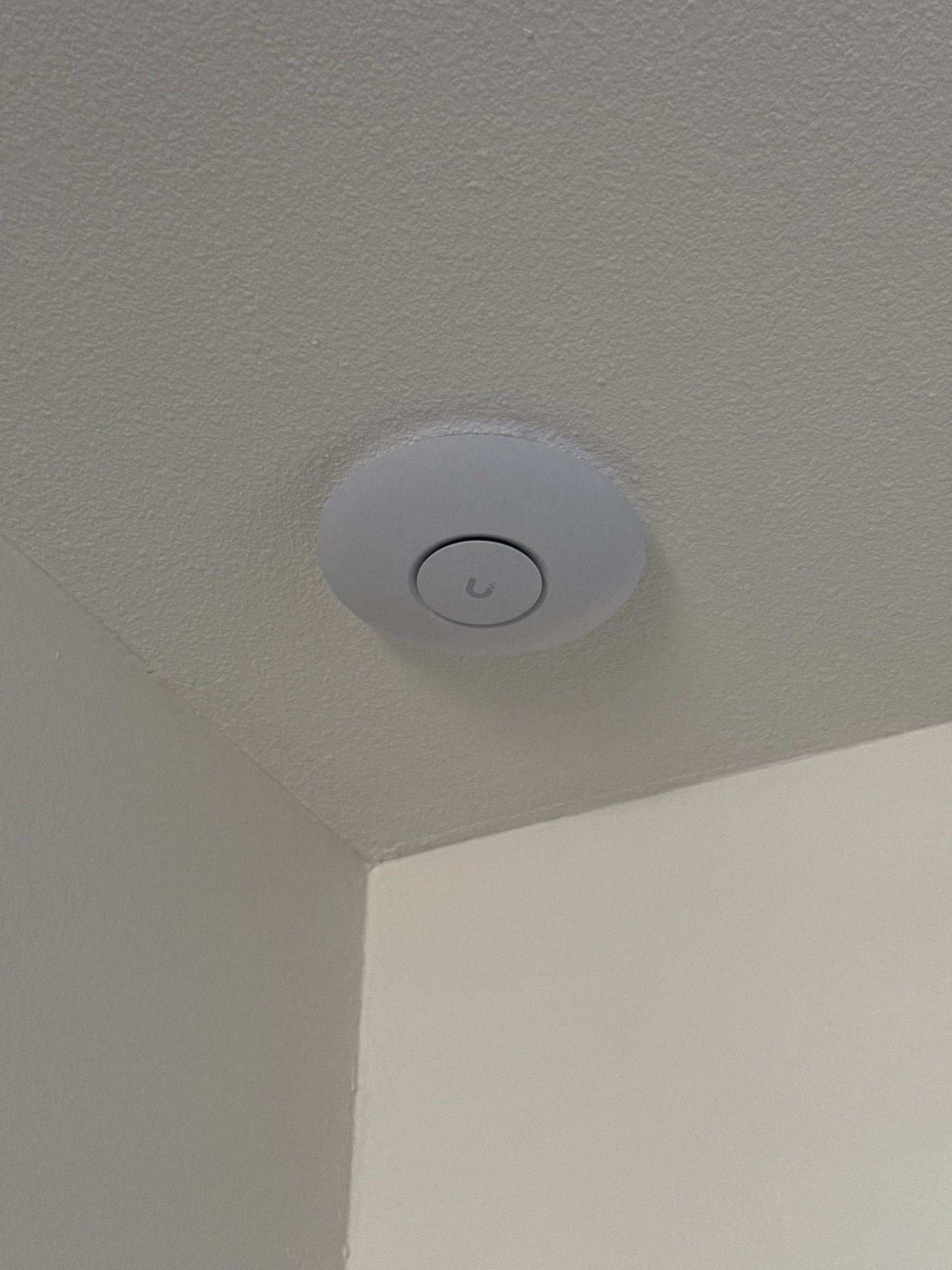 A white Ubiquiti access point is mounted on a textured ceiling corner, with the logo visible on the center cap and walls meeting below indoors.