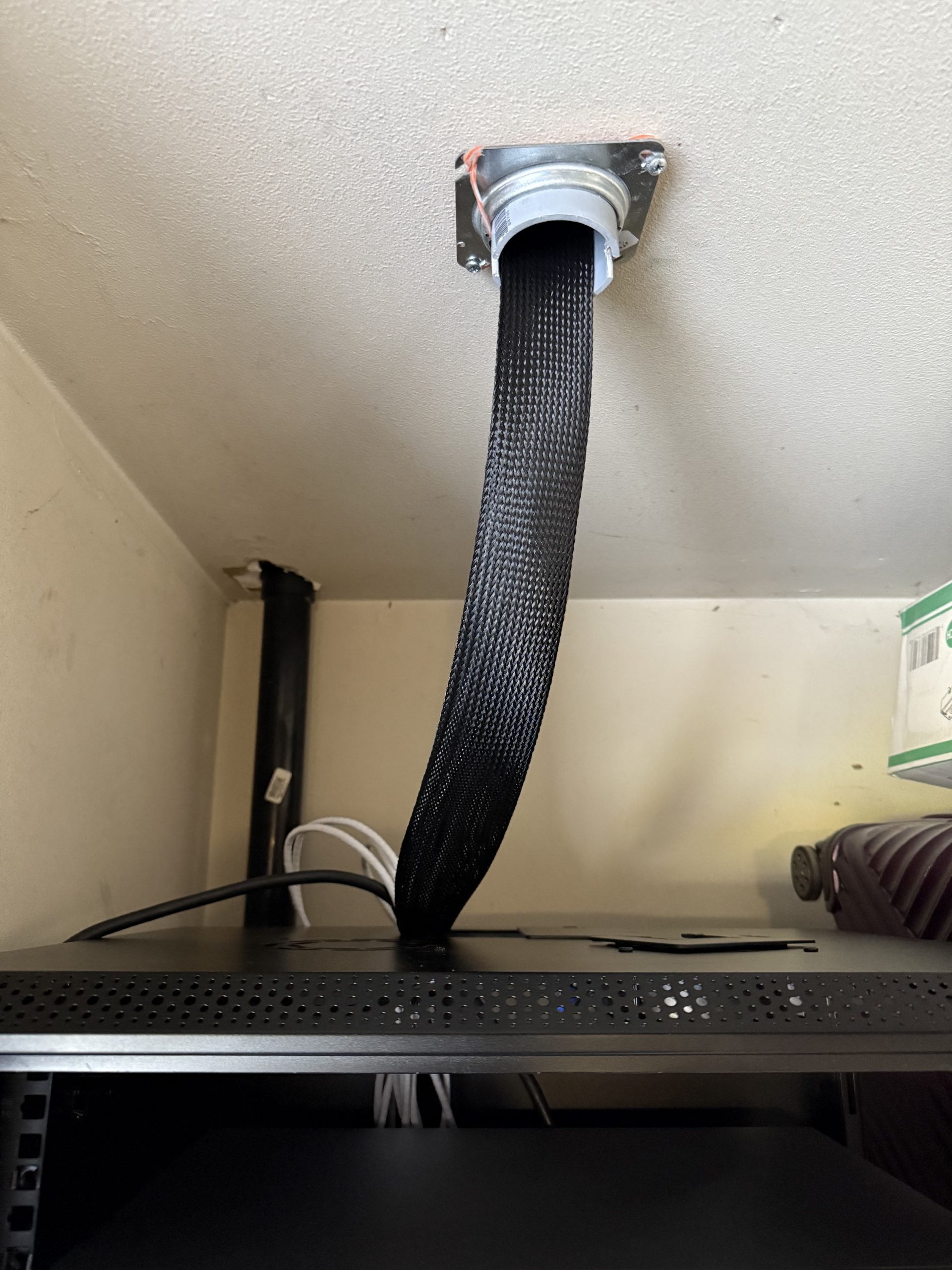 A black braided cable drops through a ceiling-mounted metal fitting into a server rack, with beige walls and exposed wiring behind it in a closet.