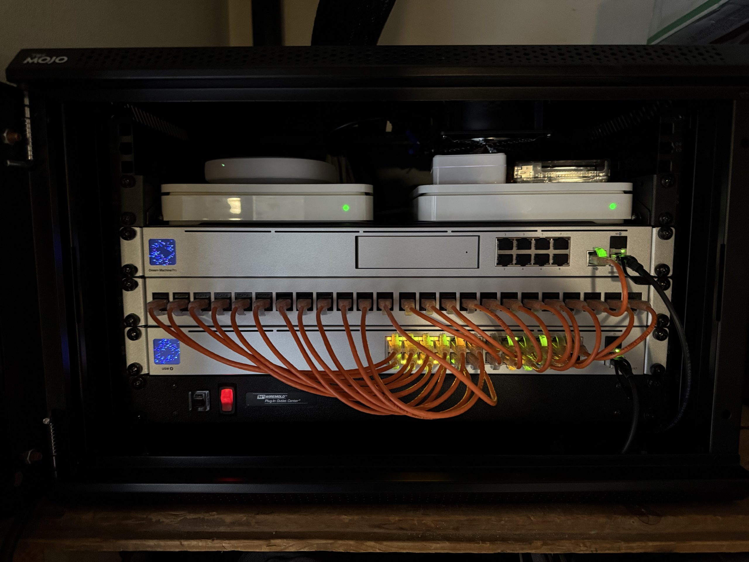 A network rack holds silver switches, two white devices, and bundled orange Ethernet cables with green status lights glowing inside the cabinet.