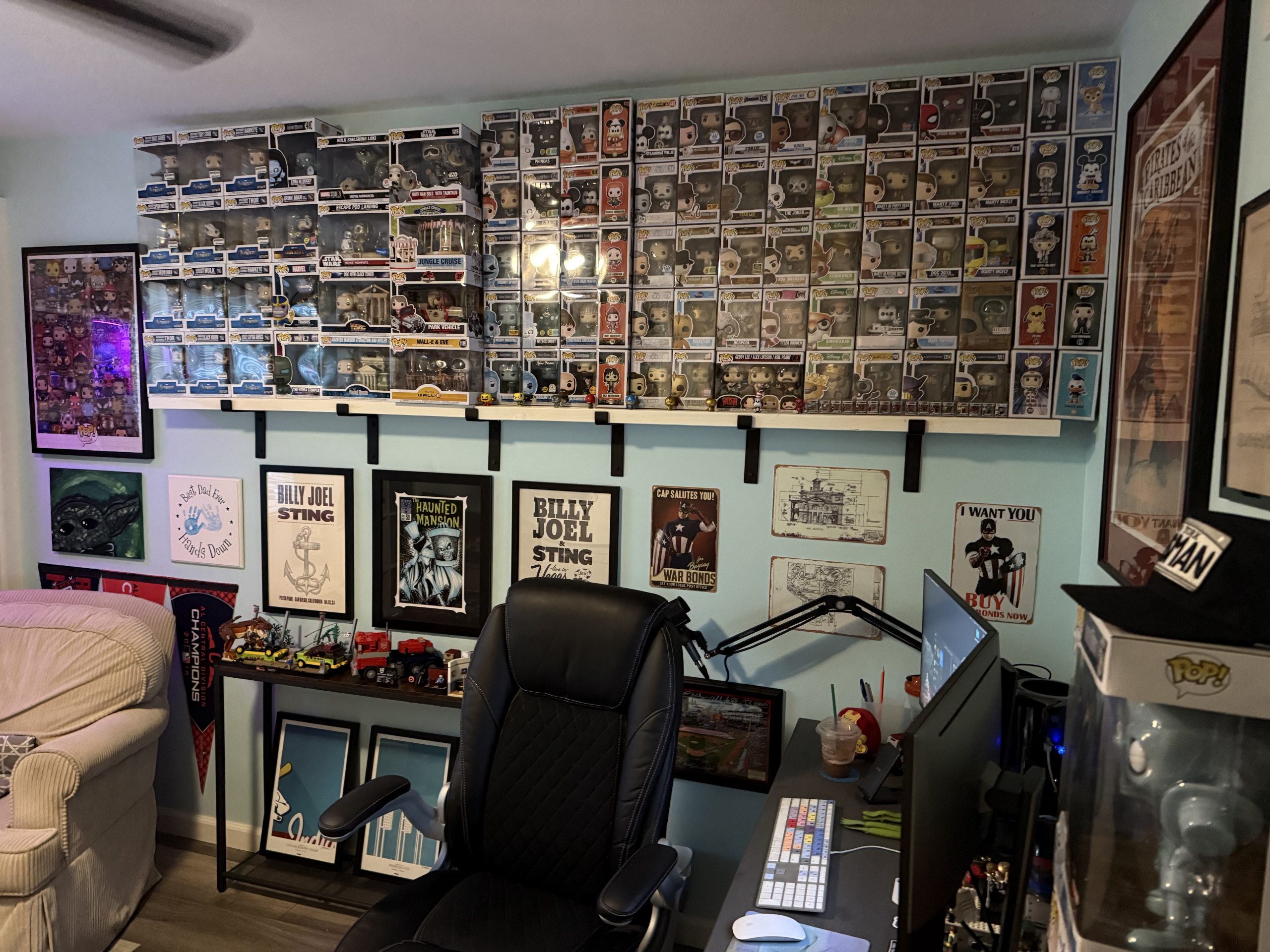 A light blue room displays wall-to-wall Funko Pop boxes, framed posters, and a black gaming chair beneath a long floating shelf and desk setup.