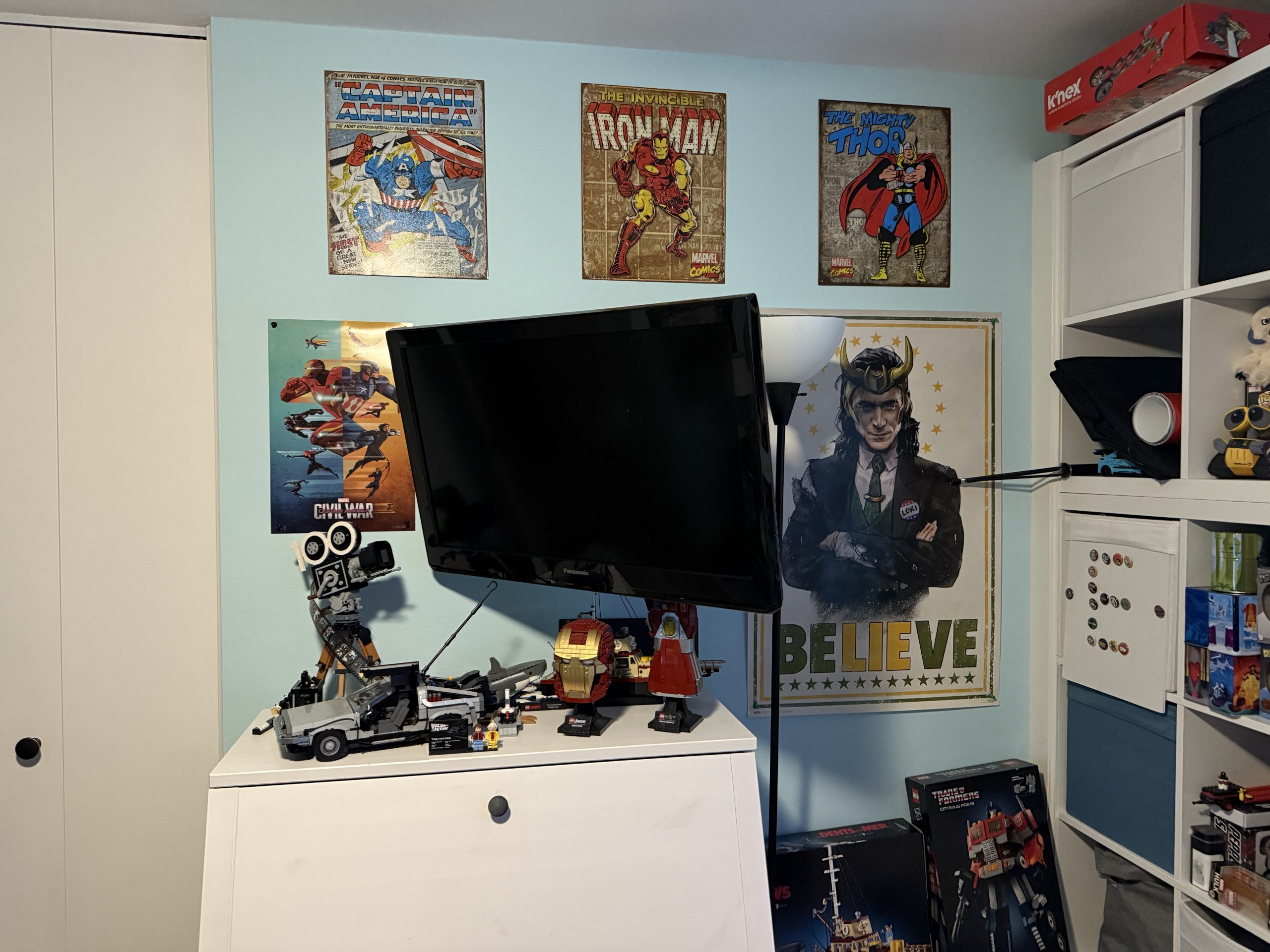 A light blue game room wall displays Captain America, Iron Man, Thor, and Loki posters above LEGO builds, a TV, and a floor lamp in a cozy corner.