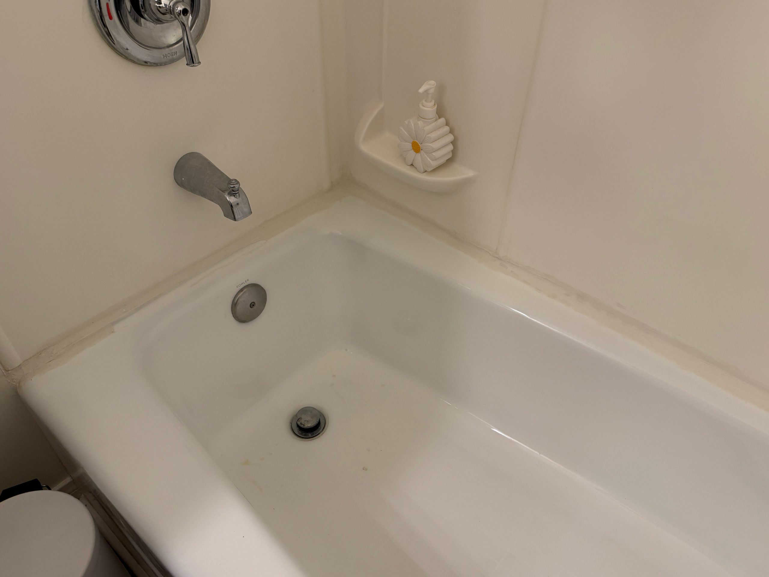 A white bathtub with chrome fixtures shows light discoloration along the caulk lines and a built-in soap dish holding a pump dispenser.