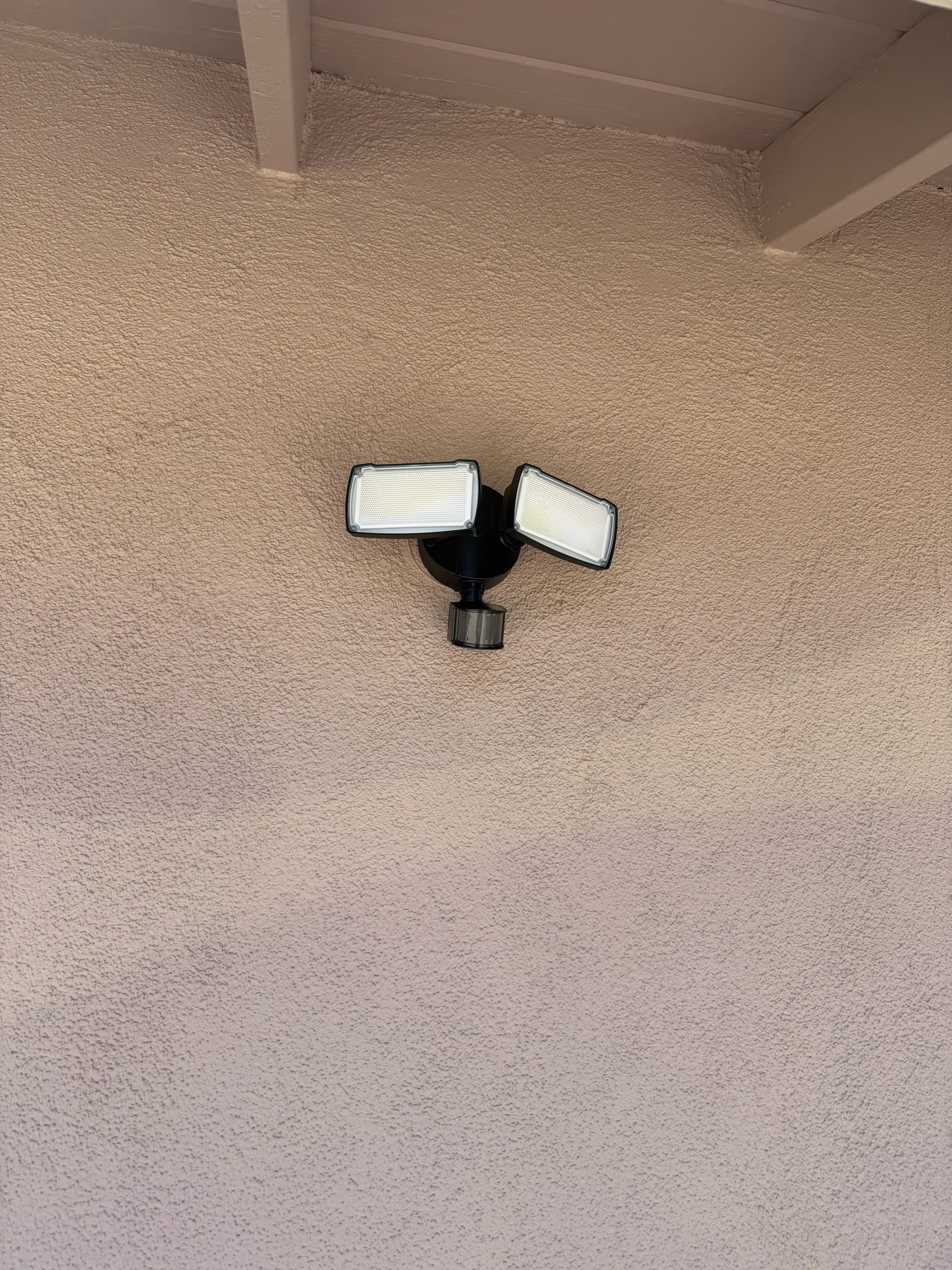 A black dual-head motion light is mounted on a beige stucco exterior wall beneath the roof eaves, with both lamp heads angled outward and downward.