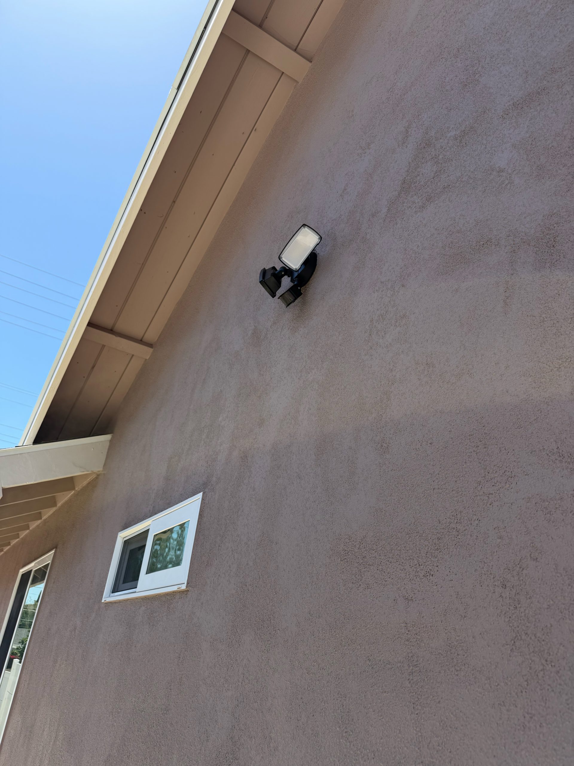 A black motion light is mounted on a stucco exterior wall beneath the eaves, with blue sky above and a white-framed window below on the side of the house.