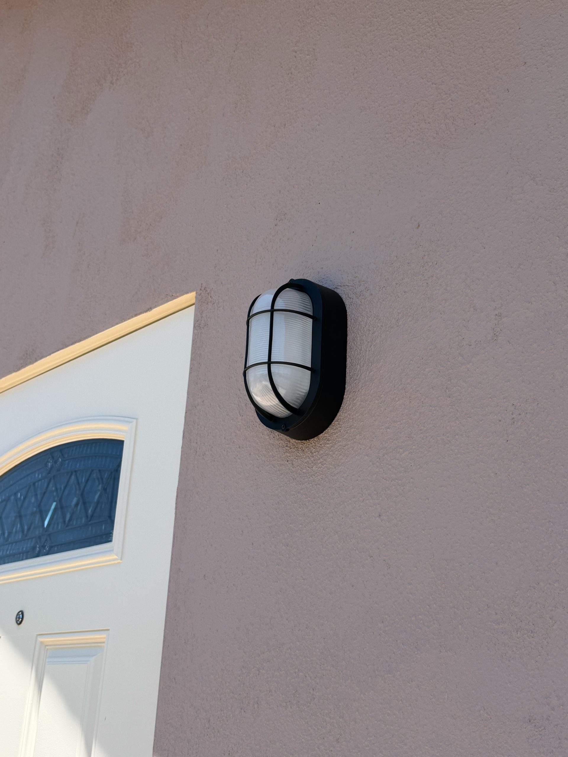 A black-caged exterior wall light glows beside a white front door on a beige stucco wall, casting a warm accent near the entryway at dusk with soft shadows.