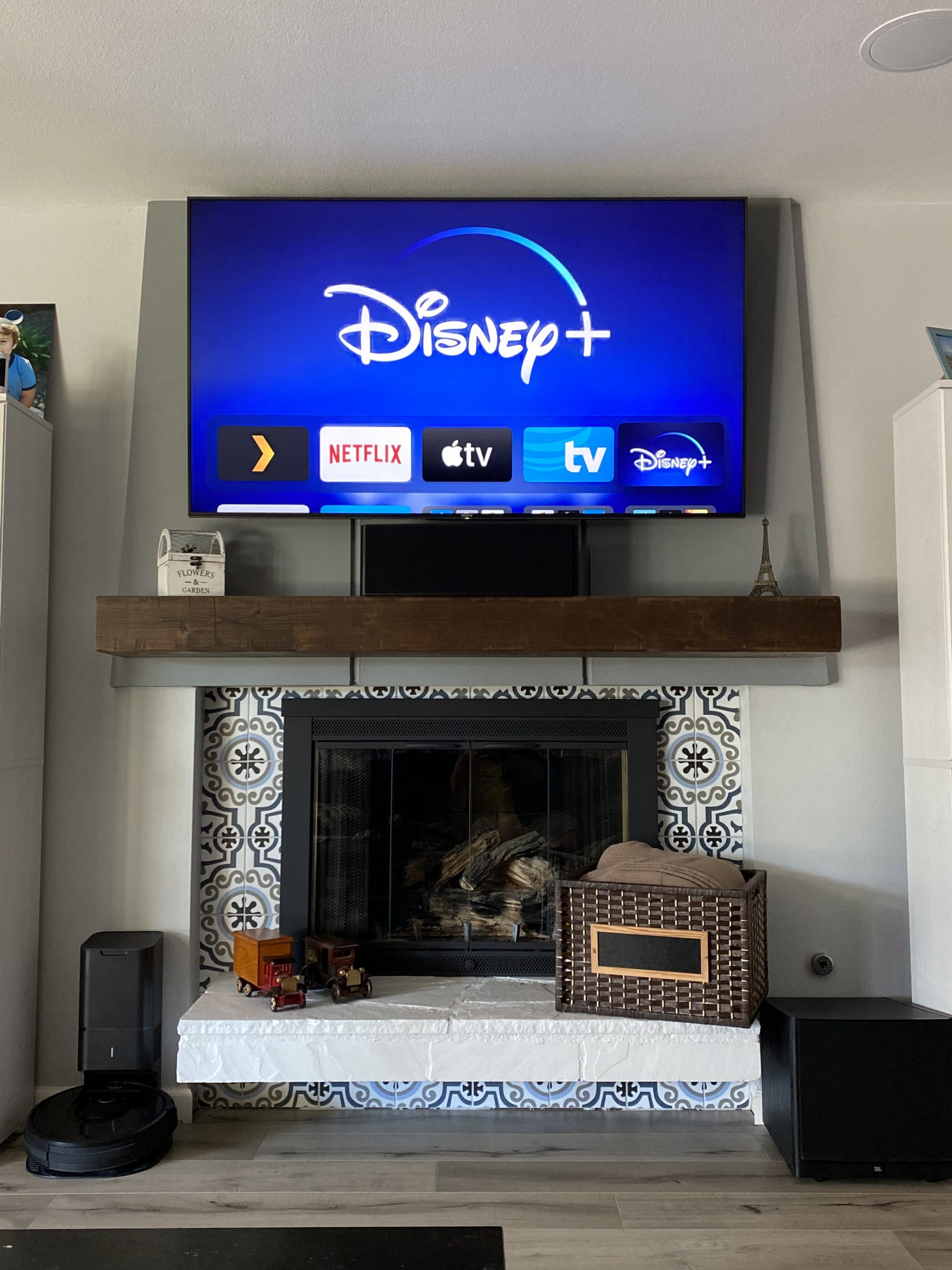 A wall-mounted TV shows Disney+ above a wooden mantel and patterned hearth tiles, with a black fireplace and small decor pieces below in a bright room.