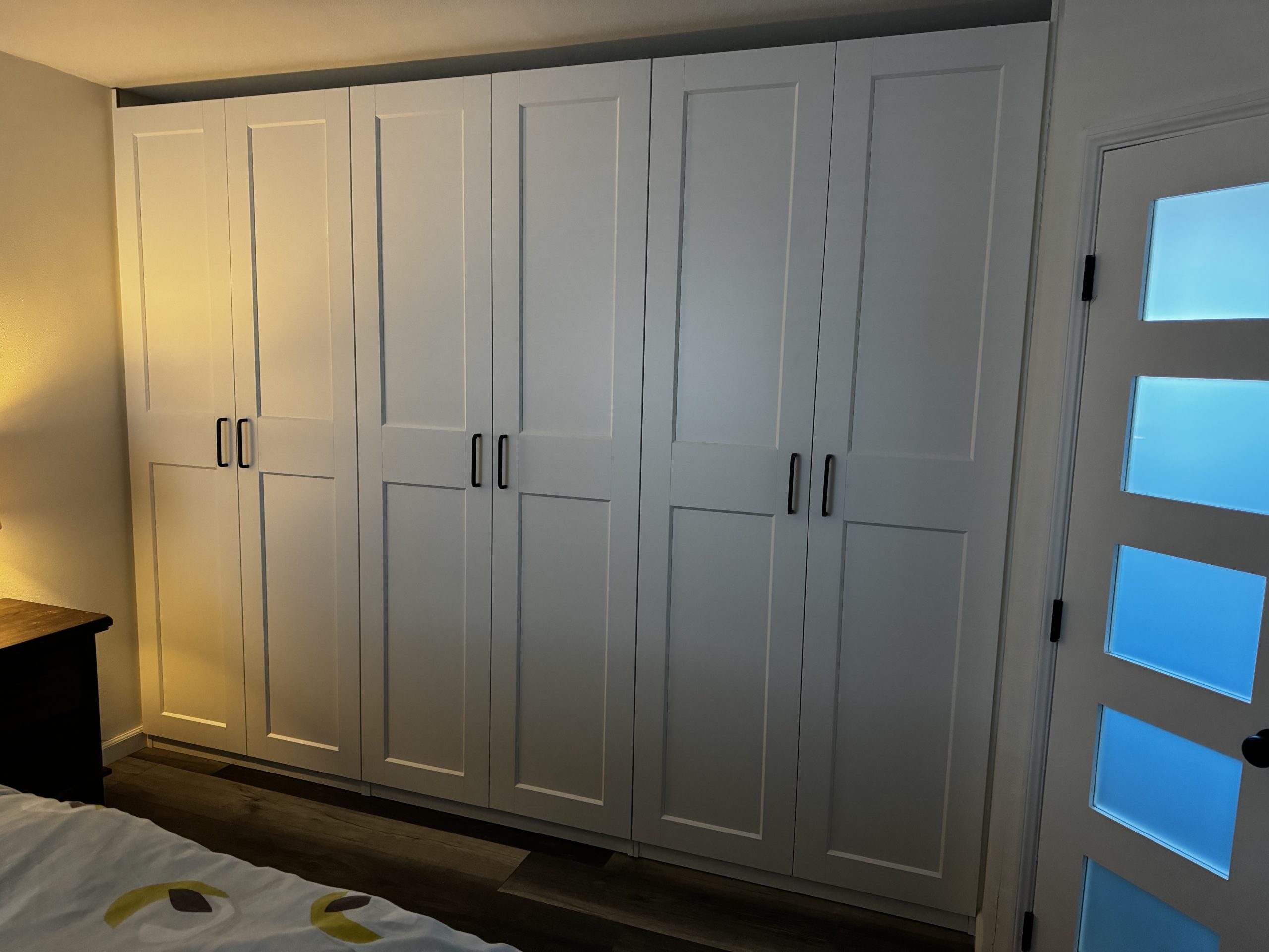 Custom white shaker closet doors with black handles line a bedroom wall beside a frosted-glass door and warm lighting over dark wood flooring.