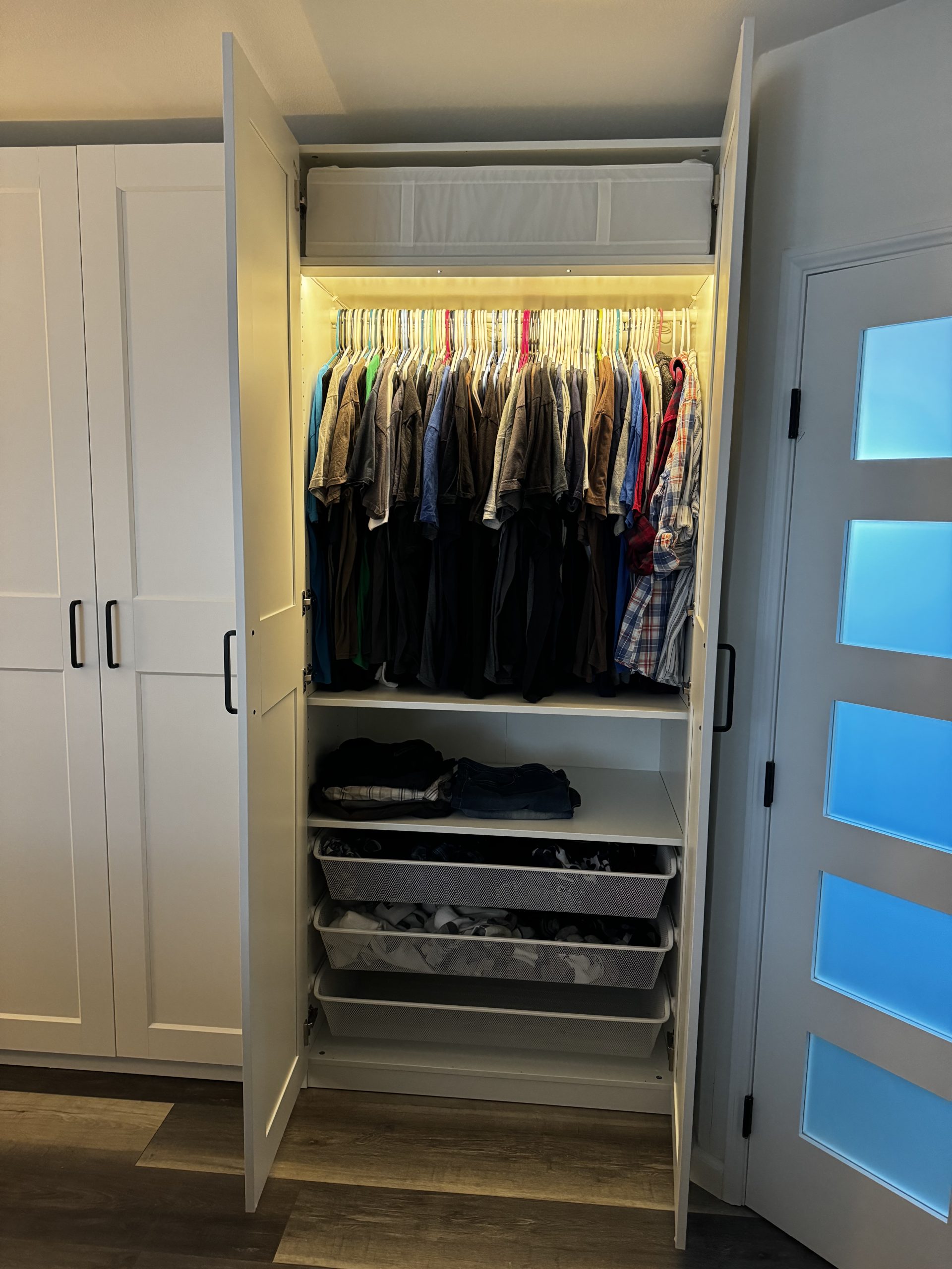 Neatly organized clothes hang inside a white built-in closet system with shelves, baskets, and warm lighting beside a frosted glass door in a bedroom.