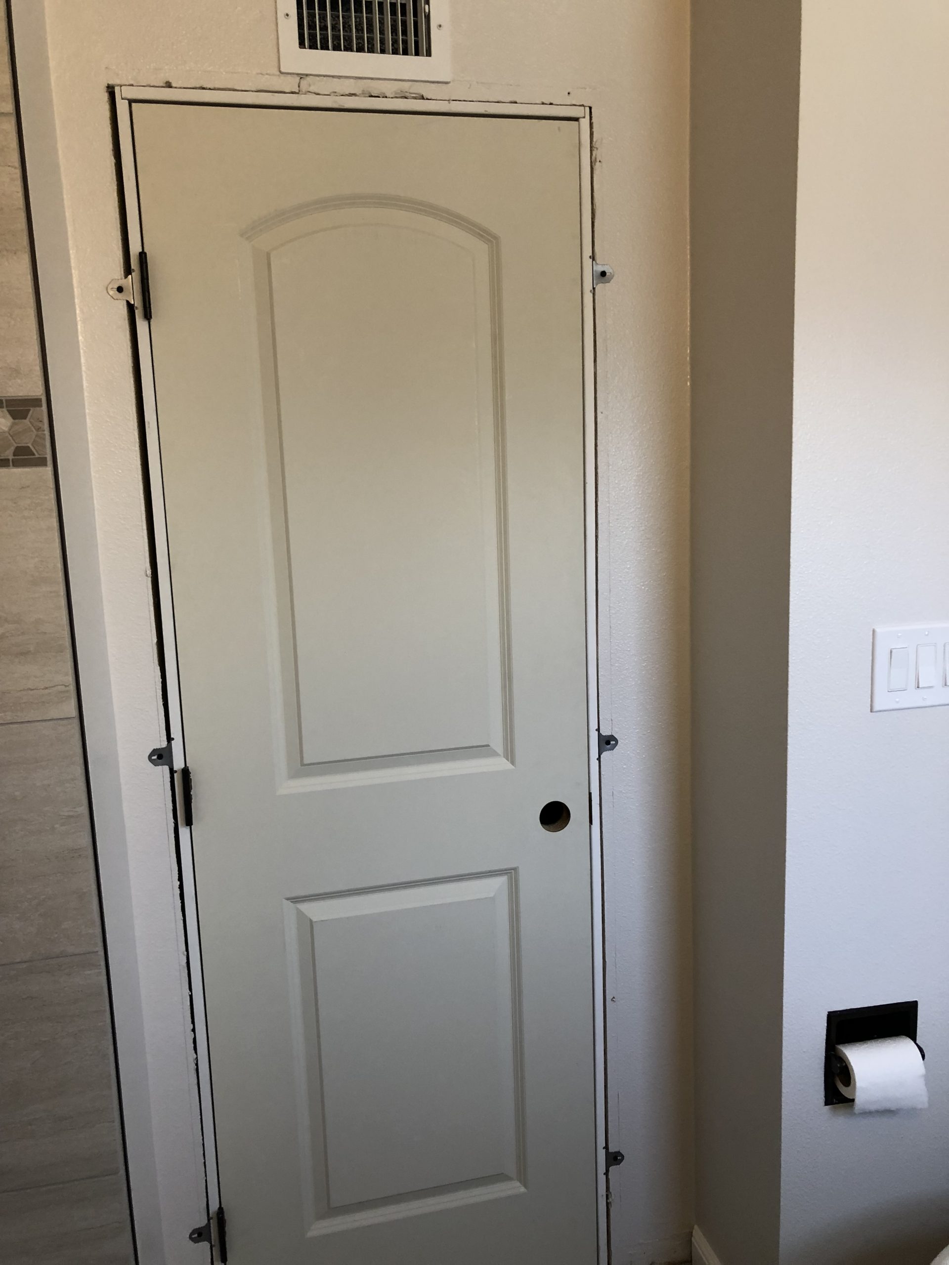 A white interior door with raised panels is installed in a bathroom doorway, with exposed hinges, a vent above, and tile walls nearby.