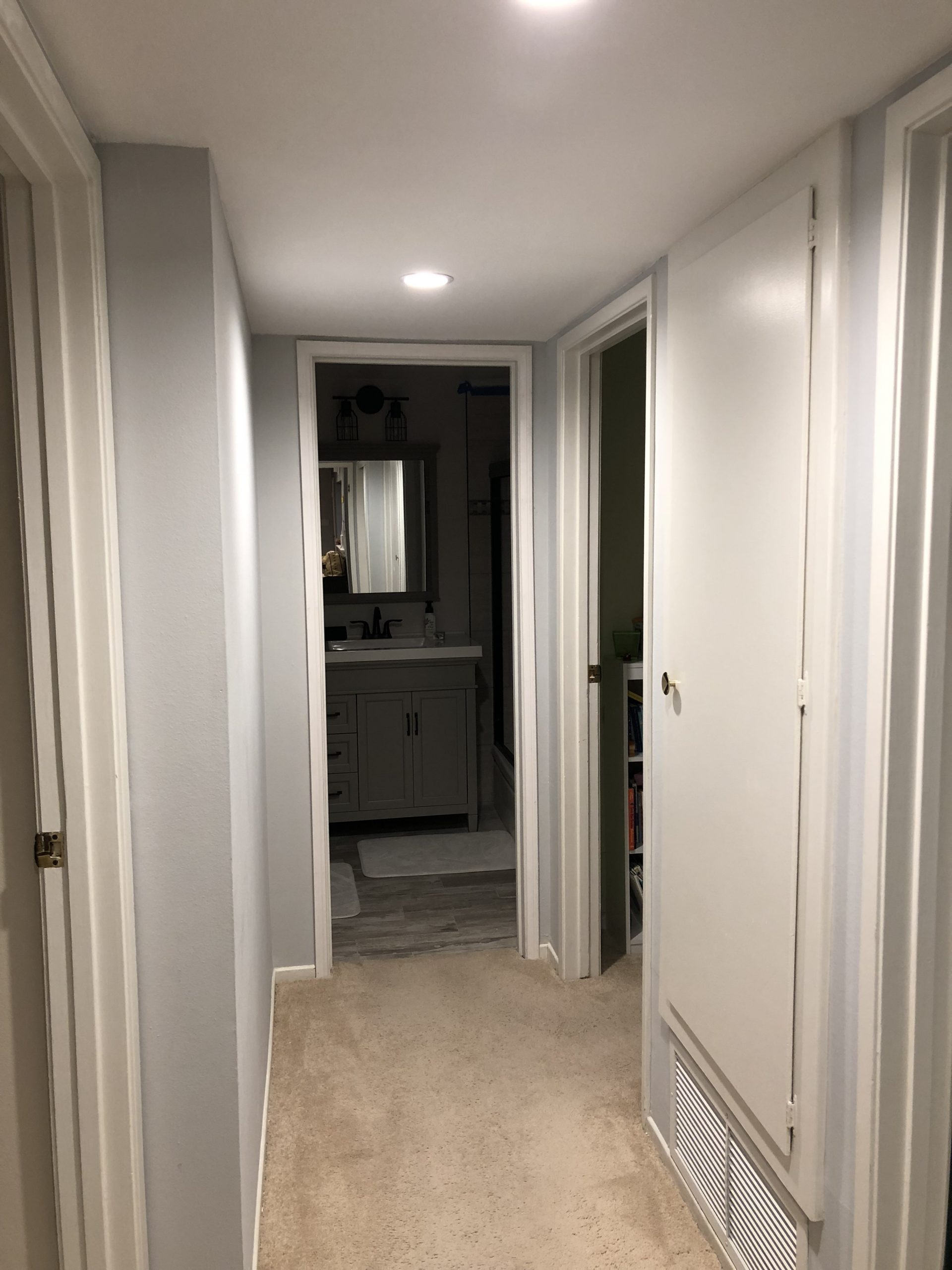 A narrow gray hallway with white trim leads past several interior doors, a recessed ceiling light, and a bathroom vanity visible ahead.