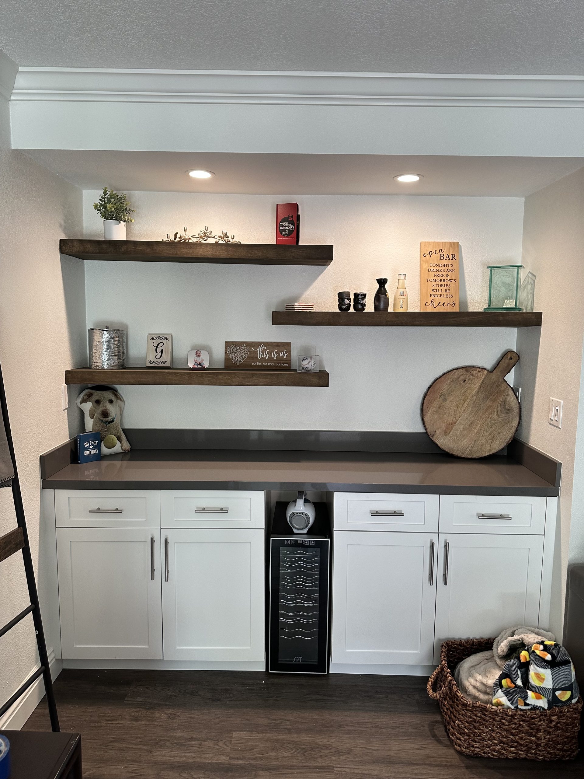 Dark floating shelves and a built-in cabinet create a stylish home bar nook with decor, recessed lights, and a wine fridge below against a white wall.