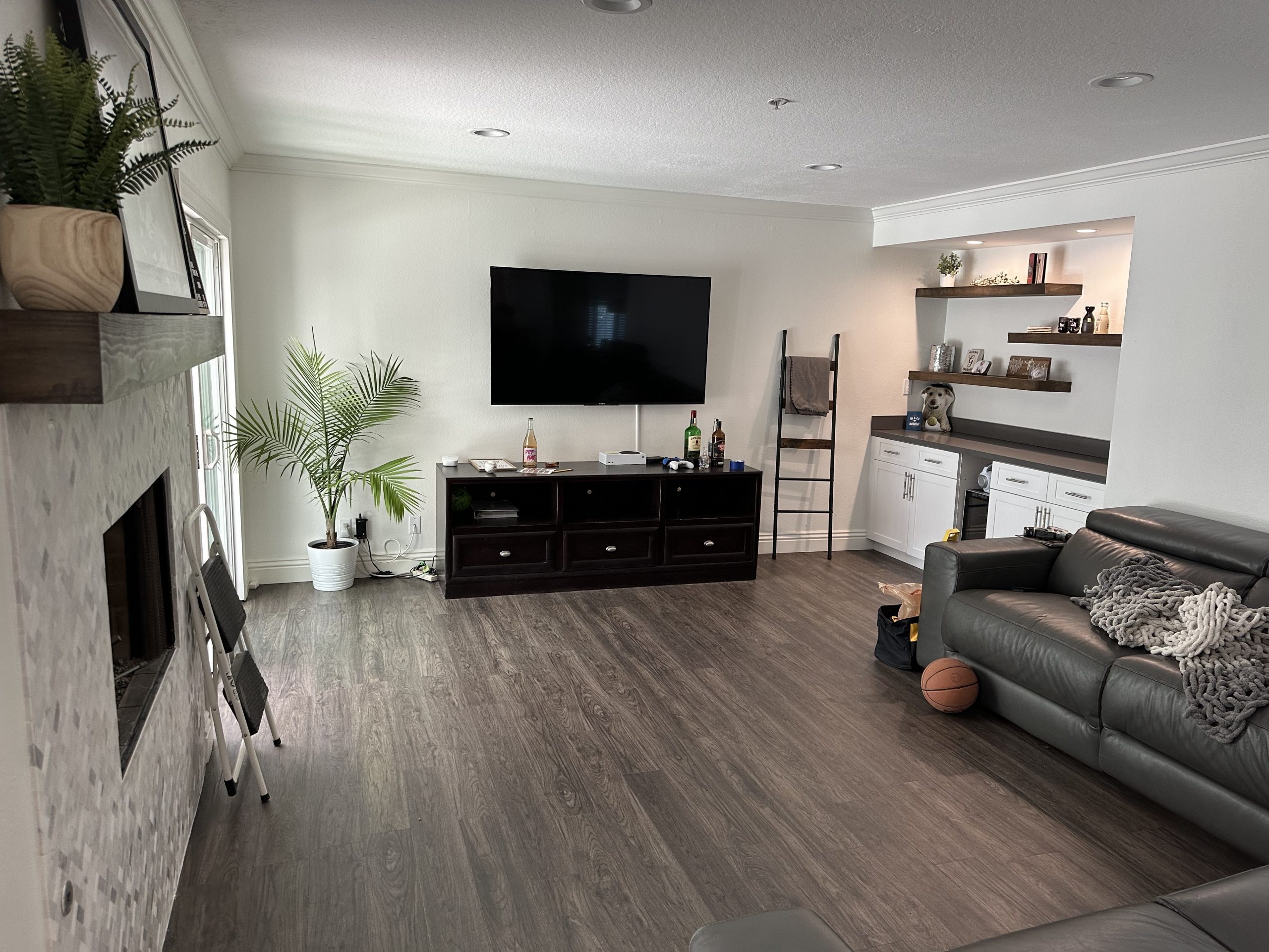 A modern living room featuring The Handy Hubs Recent Work: a wall-mounted TV, dark wood TV stand, gray sofa, potted palm, shelves with decor, basketball on the floor, and a fireplace—all set against light walls and wood flooring. , Thousand Oaks Handyman, Westlake Handyman, Oak Park Handyman, Agoura Handyman