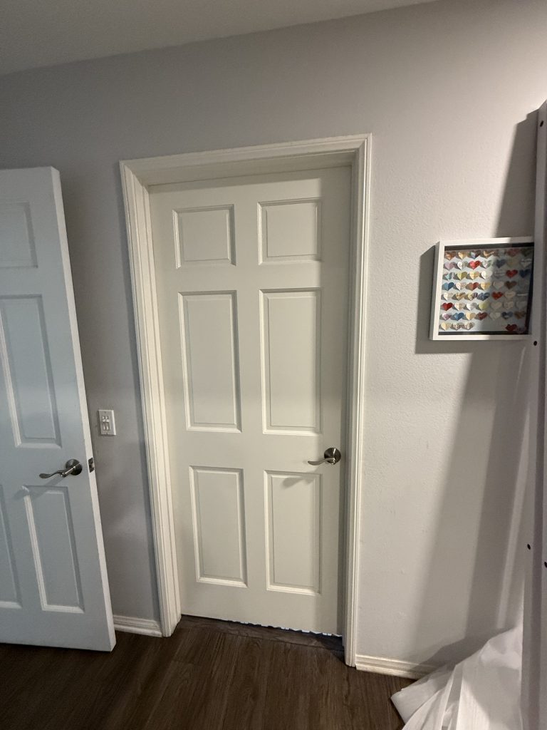An interior hallway shows a closed white paneled door beside a partly open door, with framed heart artwork, light walls, and dark wood flooring.