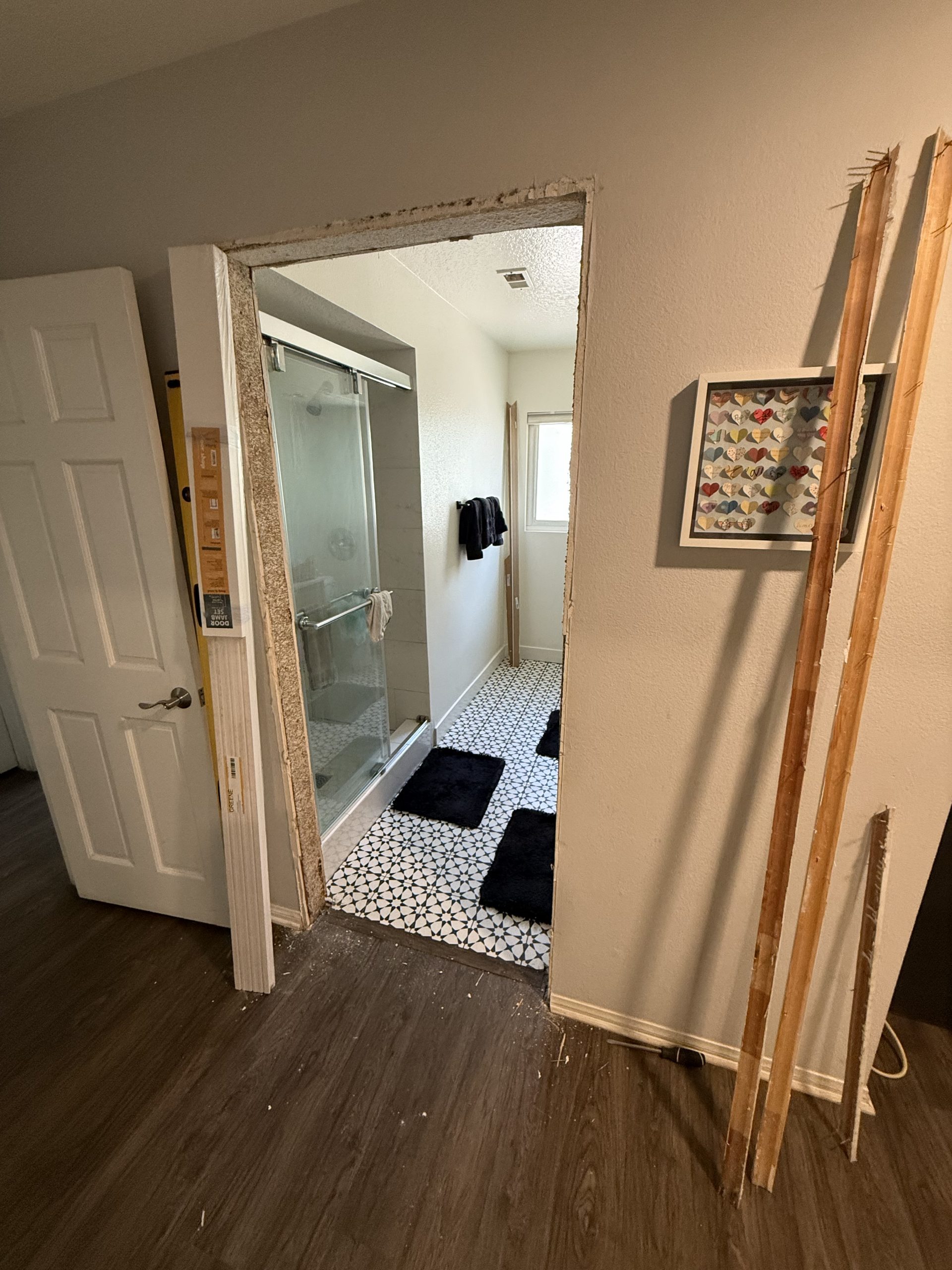 A partially installed barn door frame stands beside a bathroom with a glass shower, patterned tile floor, and loose trim pieces leaning nearby.
