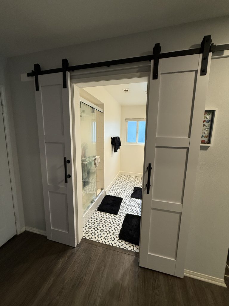 A modern bathroom from The Handy Hubs Recent Work features white sliding barn doors, patterned tile floor, black bath mats, a glass shower enclosure, white walls, and a small window letting in natural light. , Thousand Oaks Handyman, Westlake Handyman, Oak Park Handyman, Agoura Handyman