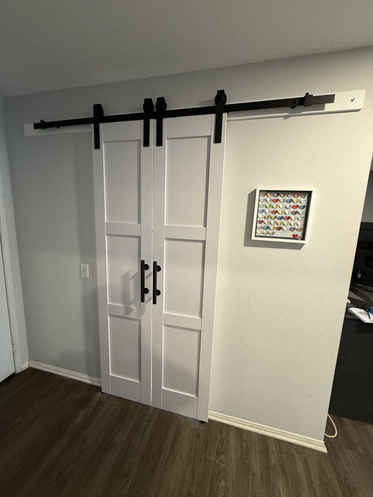 A white double sliding barn door with black hardware closes off a hallway, beside framed heart artwork and dark wood flooring in a home interior.
