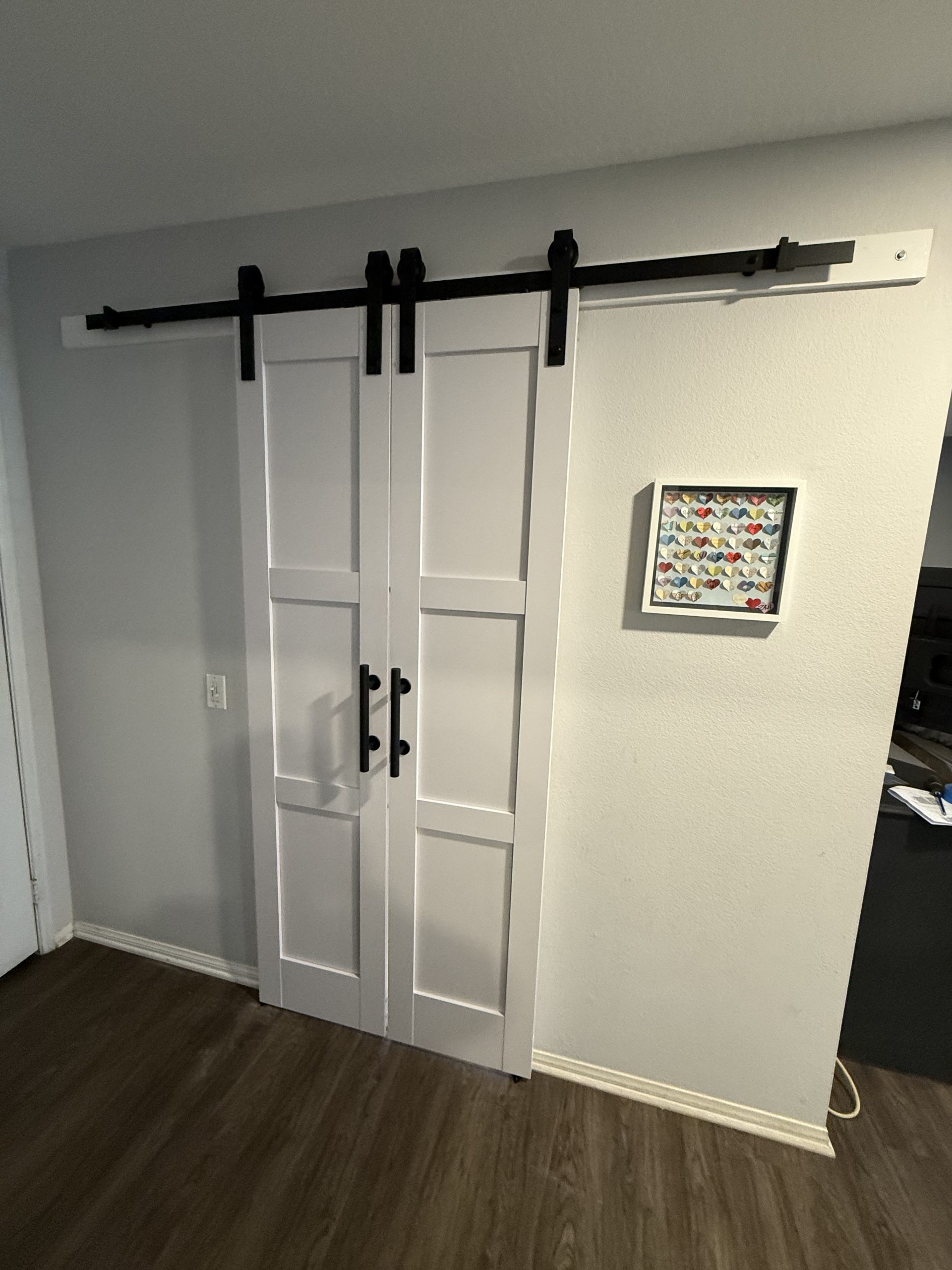 A white double sliding barn door with black hardware closes off a hallway, beside framed heart artwork and dark wood flooring in a home interior.
