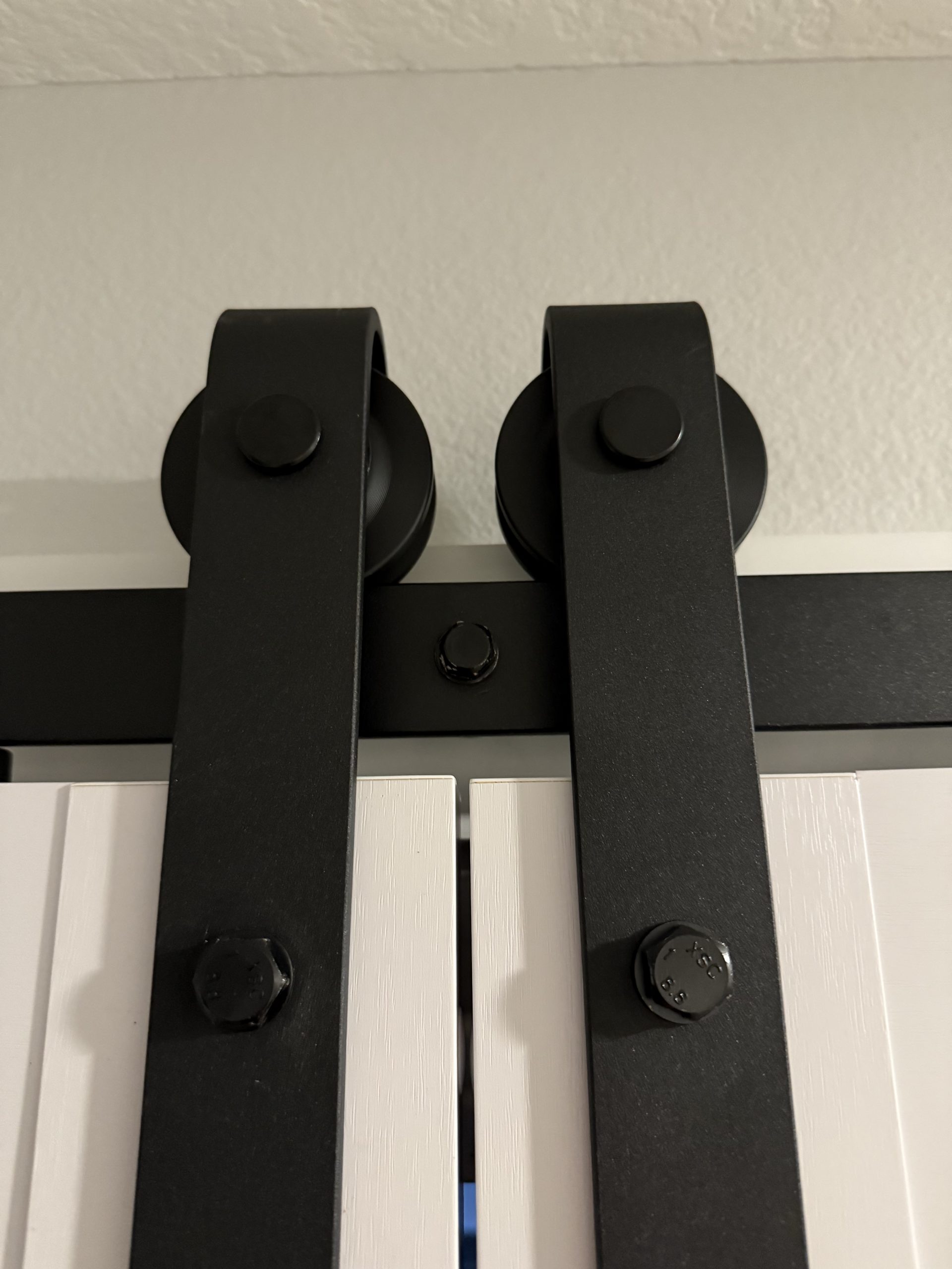 Black metal barn door hardware with twin rollers and mounting bolts secures a white sliding door against a textured wall.
