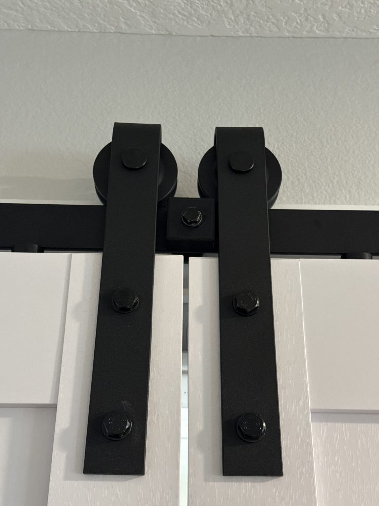Black metal barn door hardware with twin rollers and mounting bolts secures a white sliding door against a textured wall.
