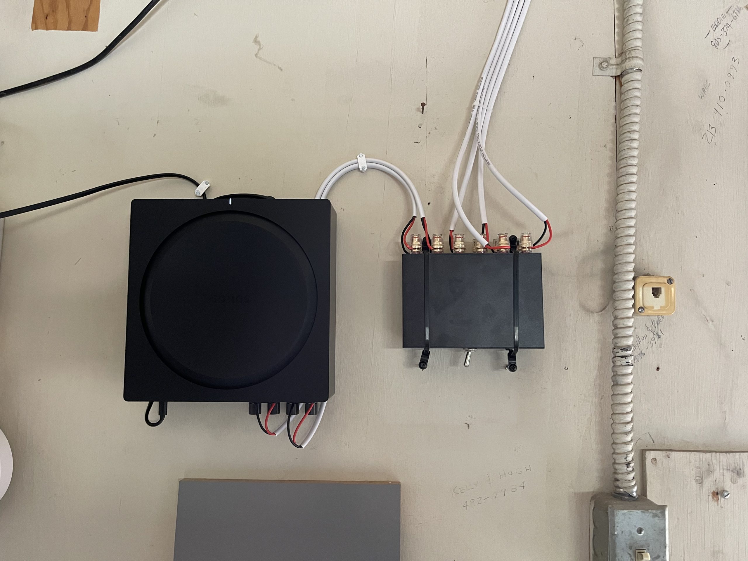 Neatly routed black and white cables connect wall-mounted audio equipment on a beige utility wall beside conduit, a network jack, and fixtures.