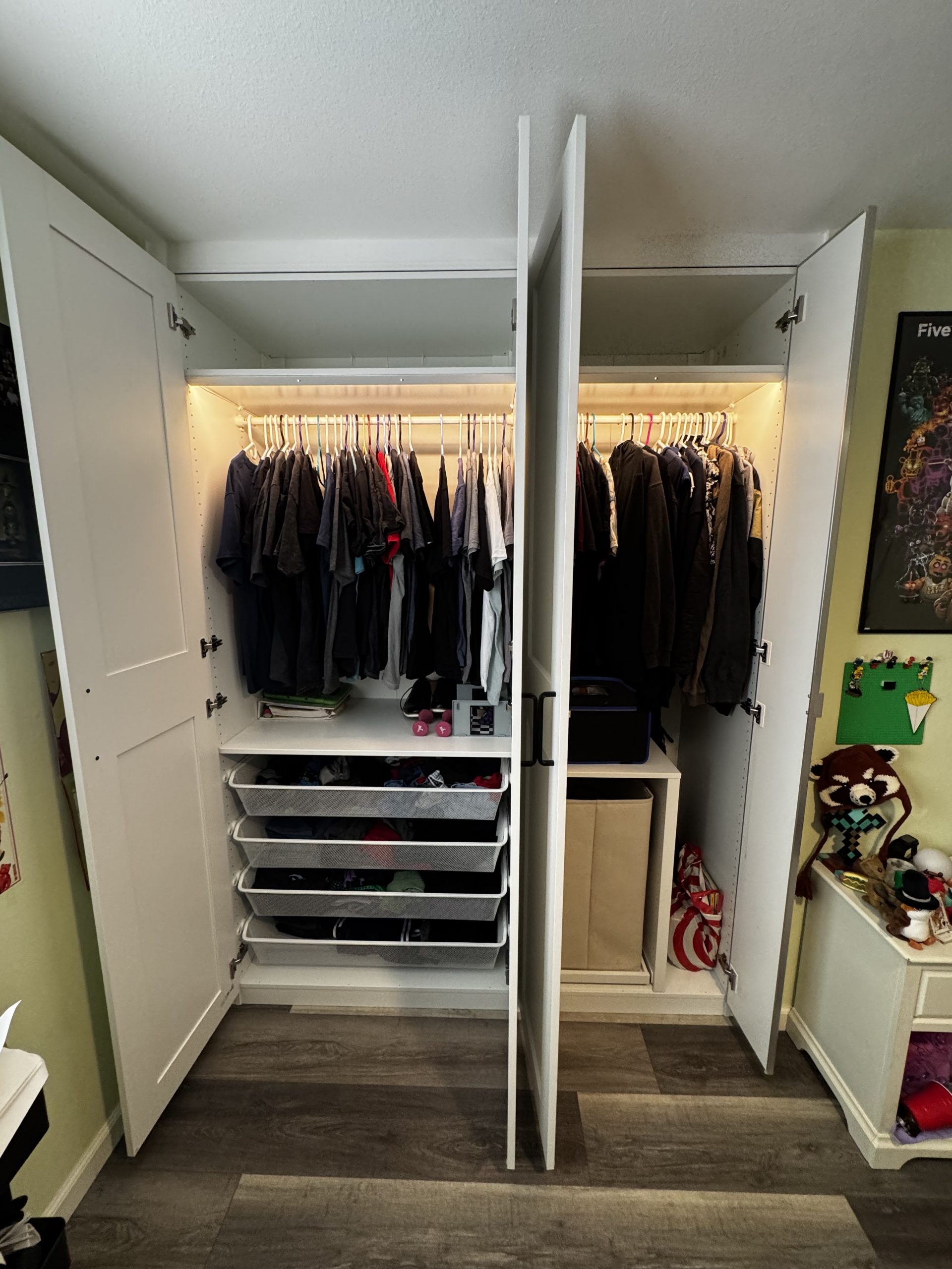 An open white wardrobe with hanging shirts, shelves with folded clothes and shoes, and some storage boxes. Warm lighting illuminates the interior—a glimpse of The Handy Hubs Recent Work. A colorful poster and stuffed toys are visible nearby. , Thousand Oaks Handyman, Westlake Handyman, Oak Park Handyman, Agoura Handyman