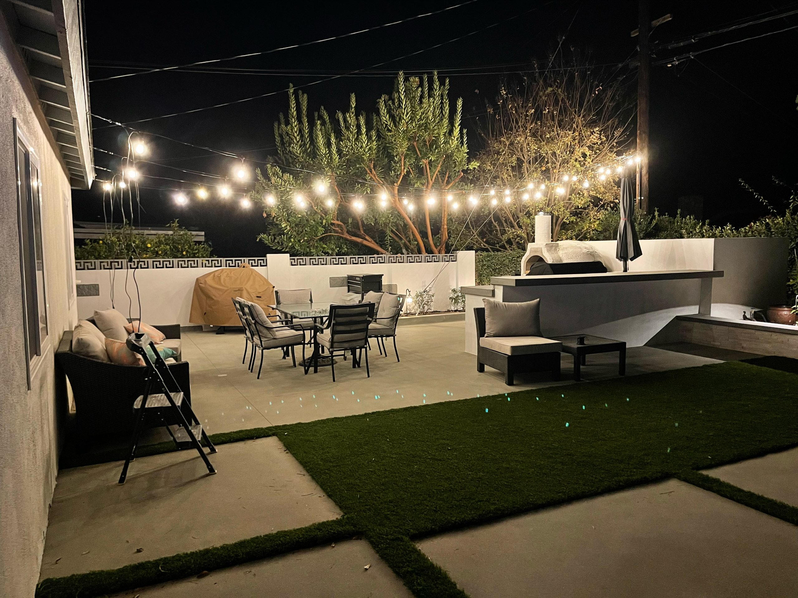 A cozy backyard patio at night, part of The Handy Hubs Recent Work, features string lights, a dining table with chairs, lounge seating, artificial grass, and a raised fire pit area, surrounded by a white wall and greenery. , Thousand Oaks Handyman, Westlake Handyman, Oak Park Handyman, Agoura Handyman