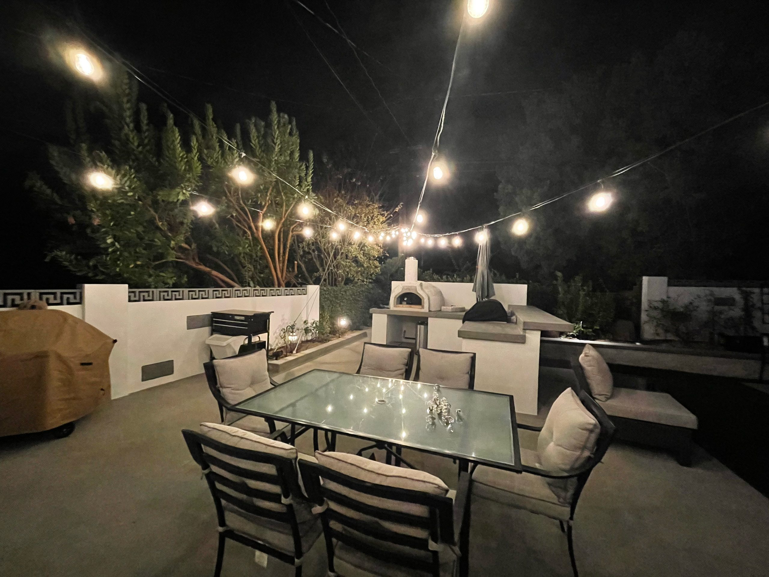 A cozy outdoor patio at night with string lights overhead, a glass-top dining table with cushioned chairs, a built-in grill, a pizza oven, and lush plants along white walls—one of The Handy Hubs Recent Work highlights. , Thousand Oaks Handyman, Westlake Handyman, Oak Park Handyman, Agoura Handyman
