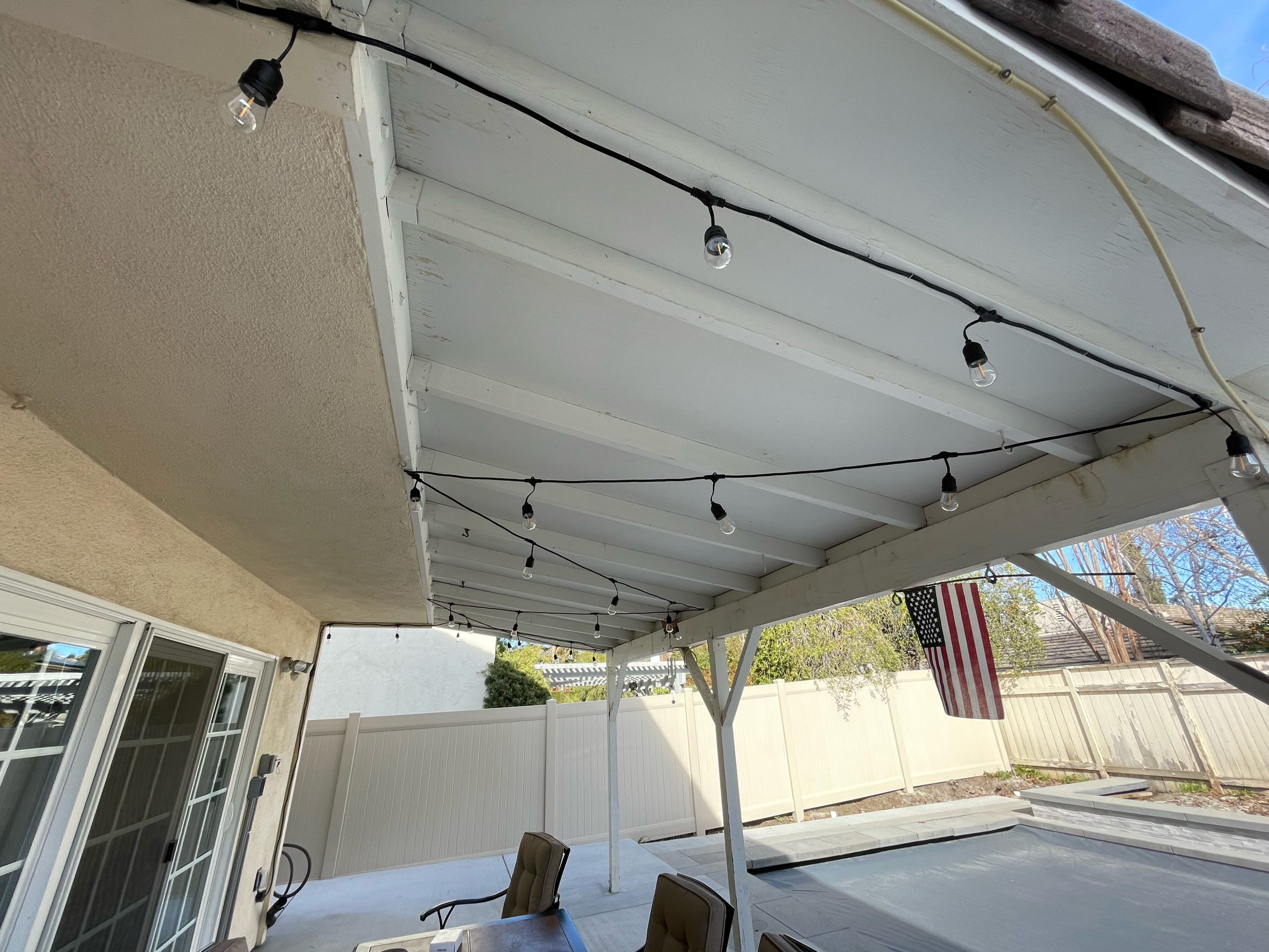 A white patio cover with string lights hangs over a patio area with outdoor chairs and a table—The Handy Hubs Recent Work. An American flag and a fenced backyard with trees are visible in the background. , Thousand Oaks Handyman, Westlake Handyman, Oak Park Handyman, Agoura Handyman