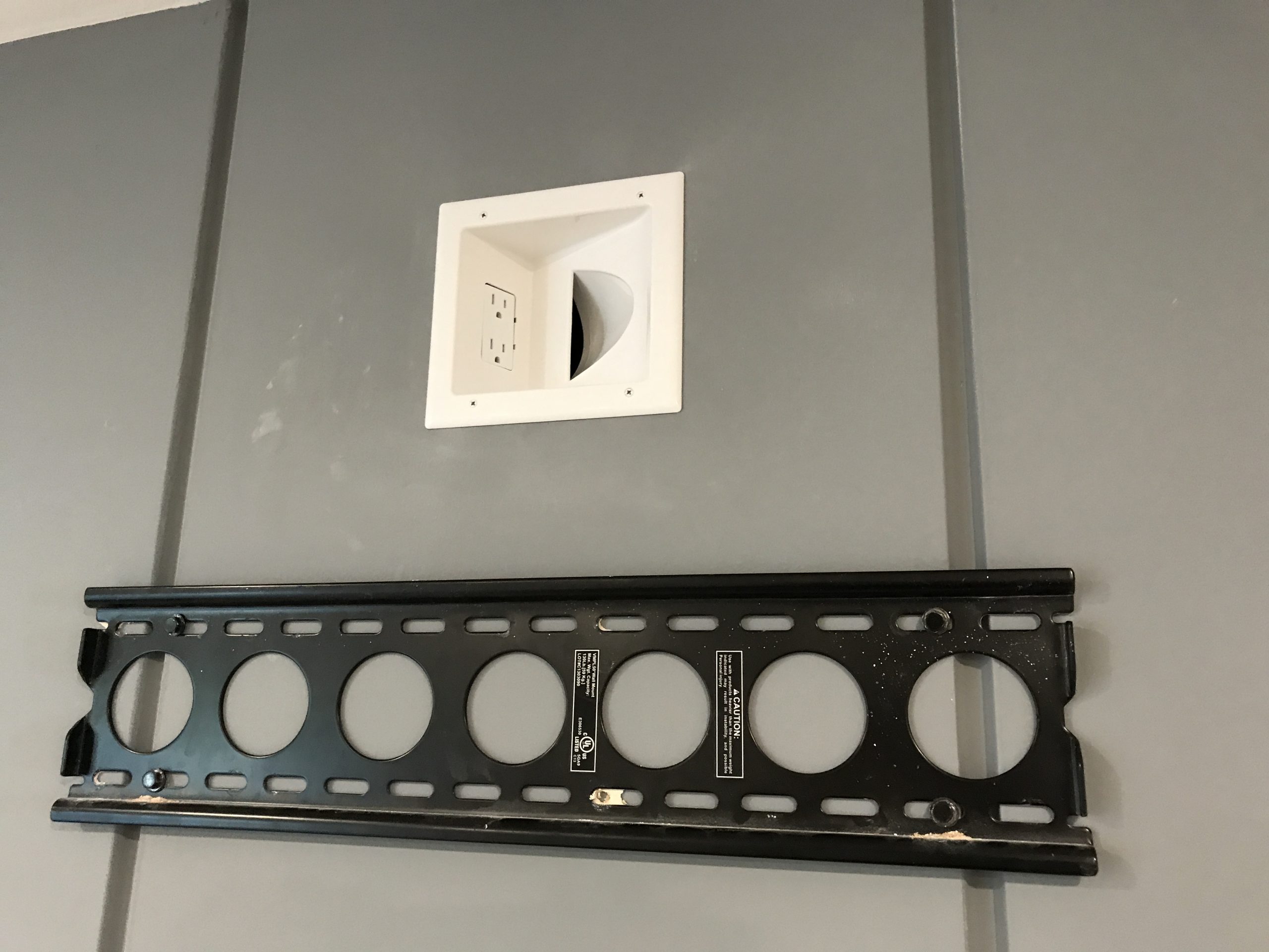 Black metal television wall mount bracket installed on a gray wall below a recessed white electrical outlet box.