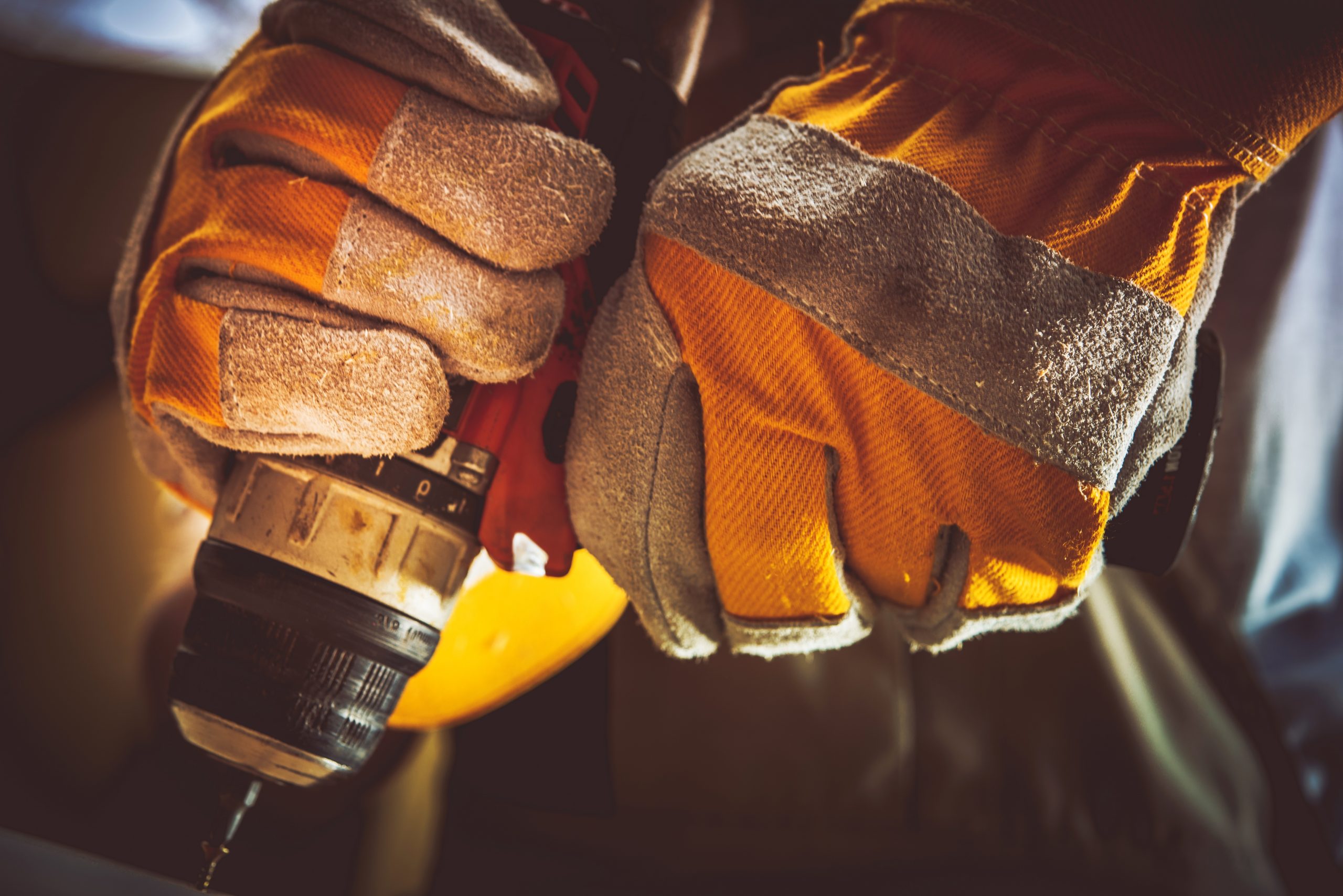 Gloved hands grip a power drill with a bit attached, showing yellow protective work gloves and a close-up view of handyman tools in action.