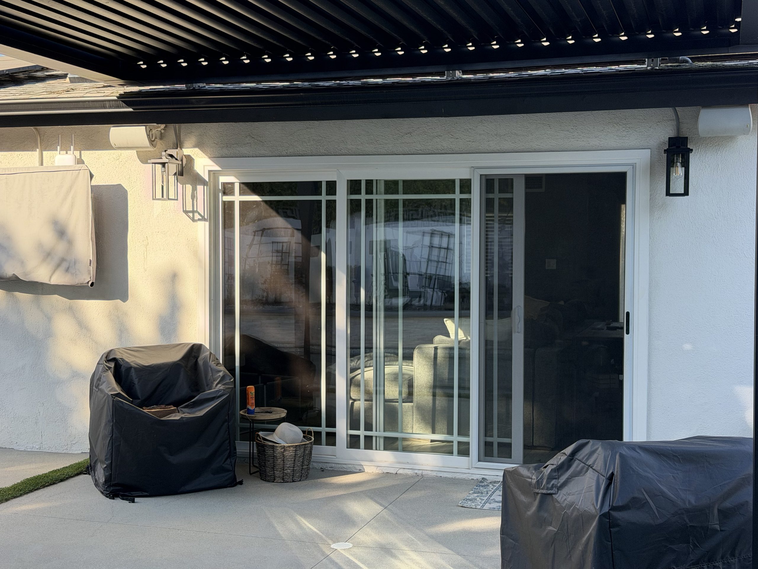Backyard patio with a sliding glass door, black pergola slats overhead, wall lanterns, and two covered grills.